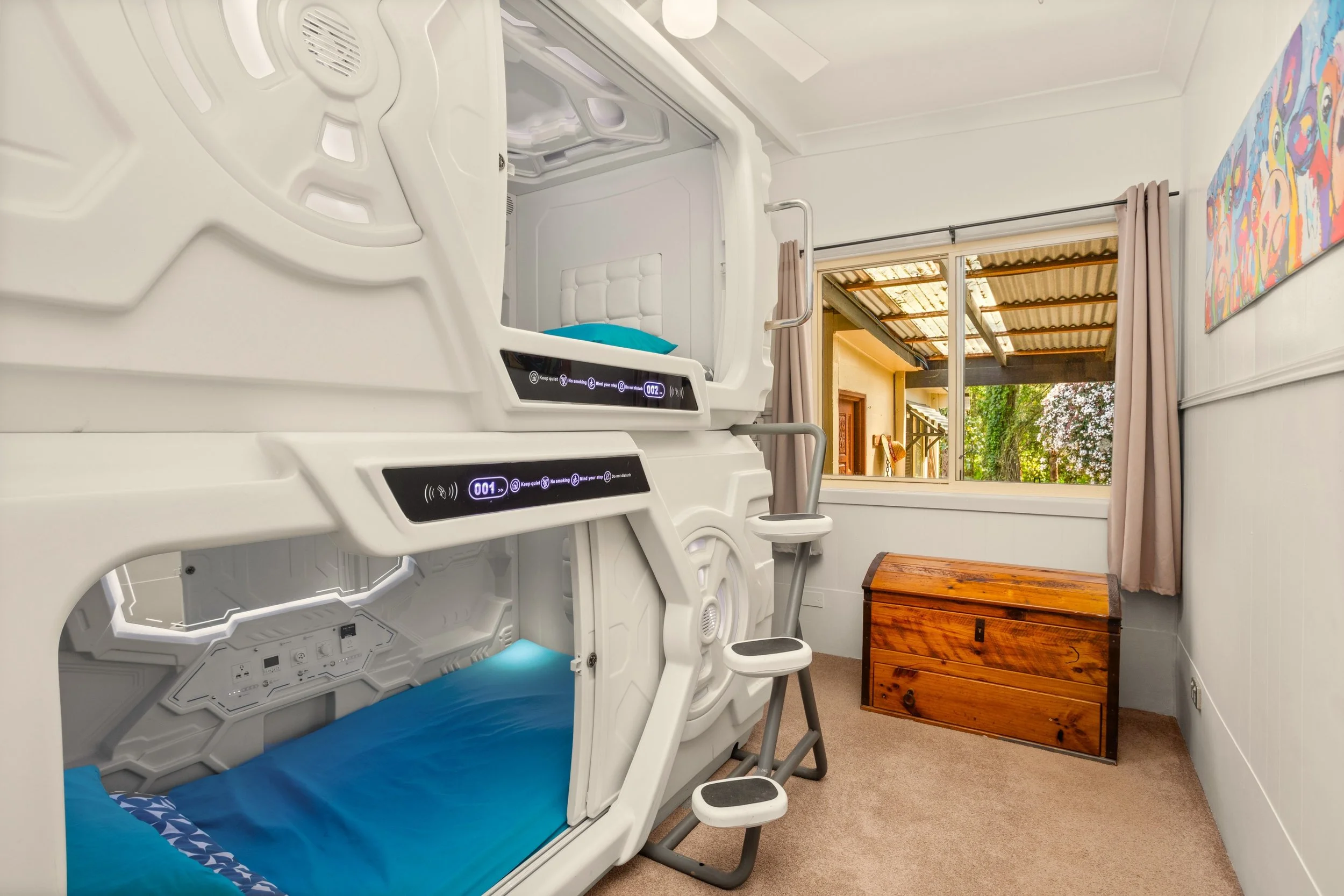 A room with two sci-fi style bunk beds with built-in digital controls, a ladder, a window with curtains showing an outdoor scene with trees, and a wooden chest at the corner.
