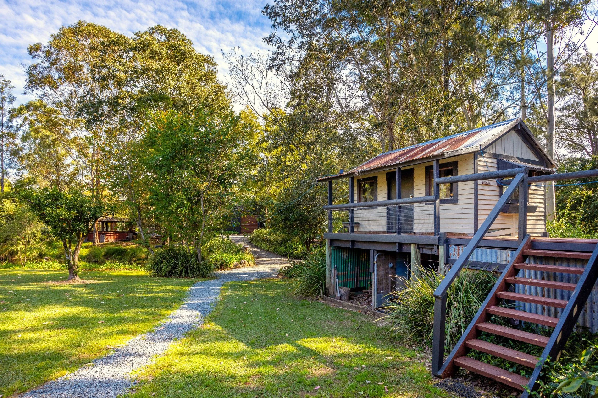 A small house on stilts with a red rusted metal roof and wooden stairs, surrounded by a lush green yard with a gravel pathway, trees, and bushes.