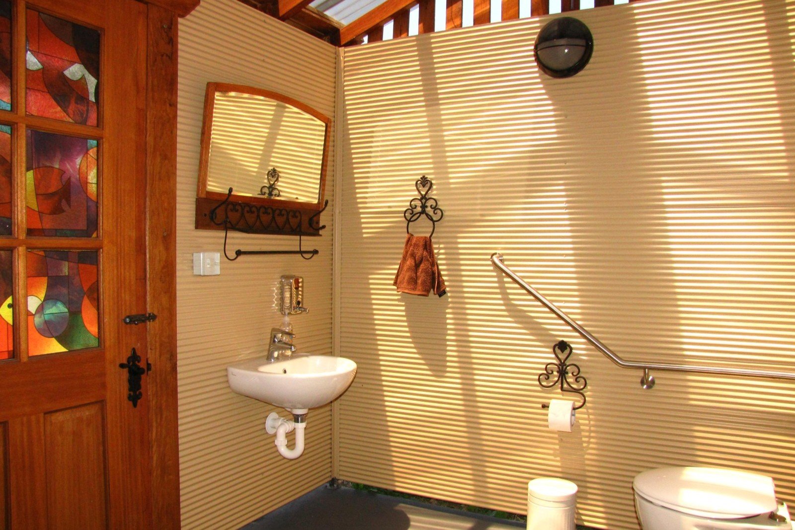 A bathroom with beige ribbed wall panels, a small white wall-mounted sink, a toilet, a mirror with a wooden frame, a towel rack with a brown towel, a toilet paper holder with a decorative metal design, a trash can, a black wall light, and a wooden do