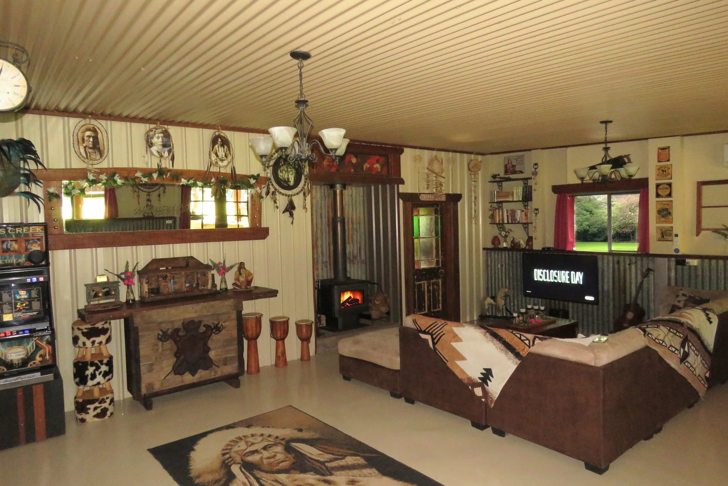 Living room with wood-paneled ceiling, vintage decor, a couch with Native American-style blankets, a fireplace, and a television displaying the words 'Disclosure Day'.