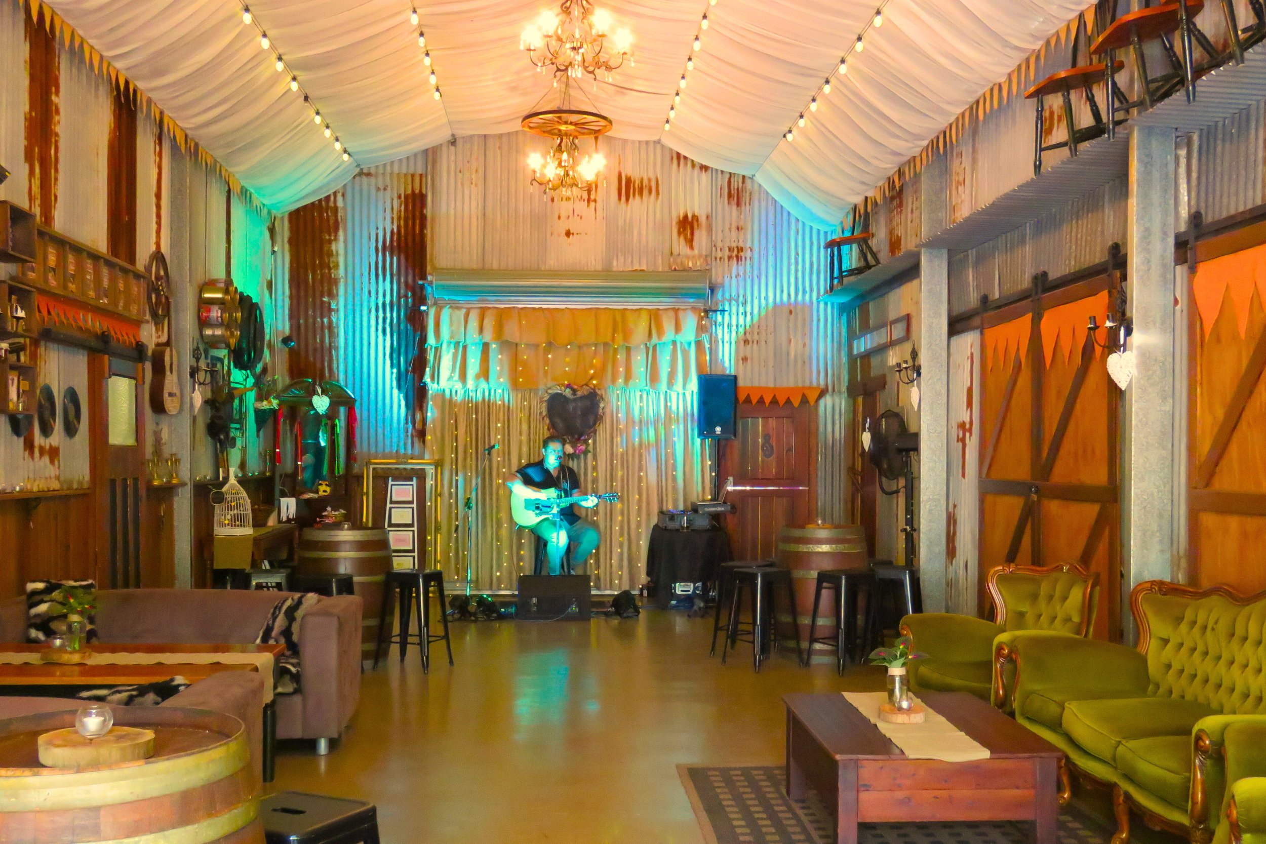 Interior of a rustic bar or restaurant with a small stage featuring a musician playing guitar, surrounded by vintage decor, wooden furniture, and string lights.