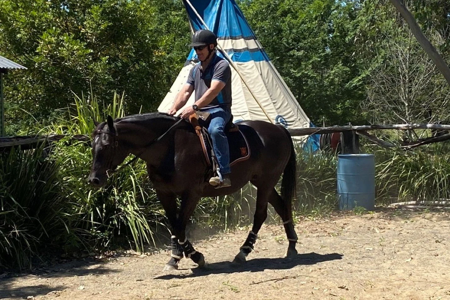 A man riding a brown horse on a dirt path with a teepee tent and green trees in the background.