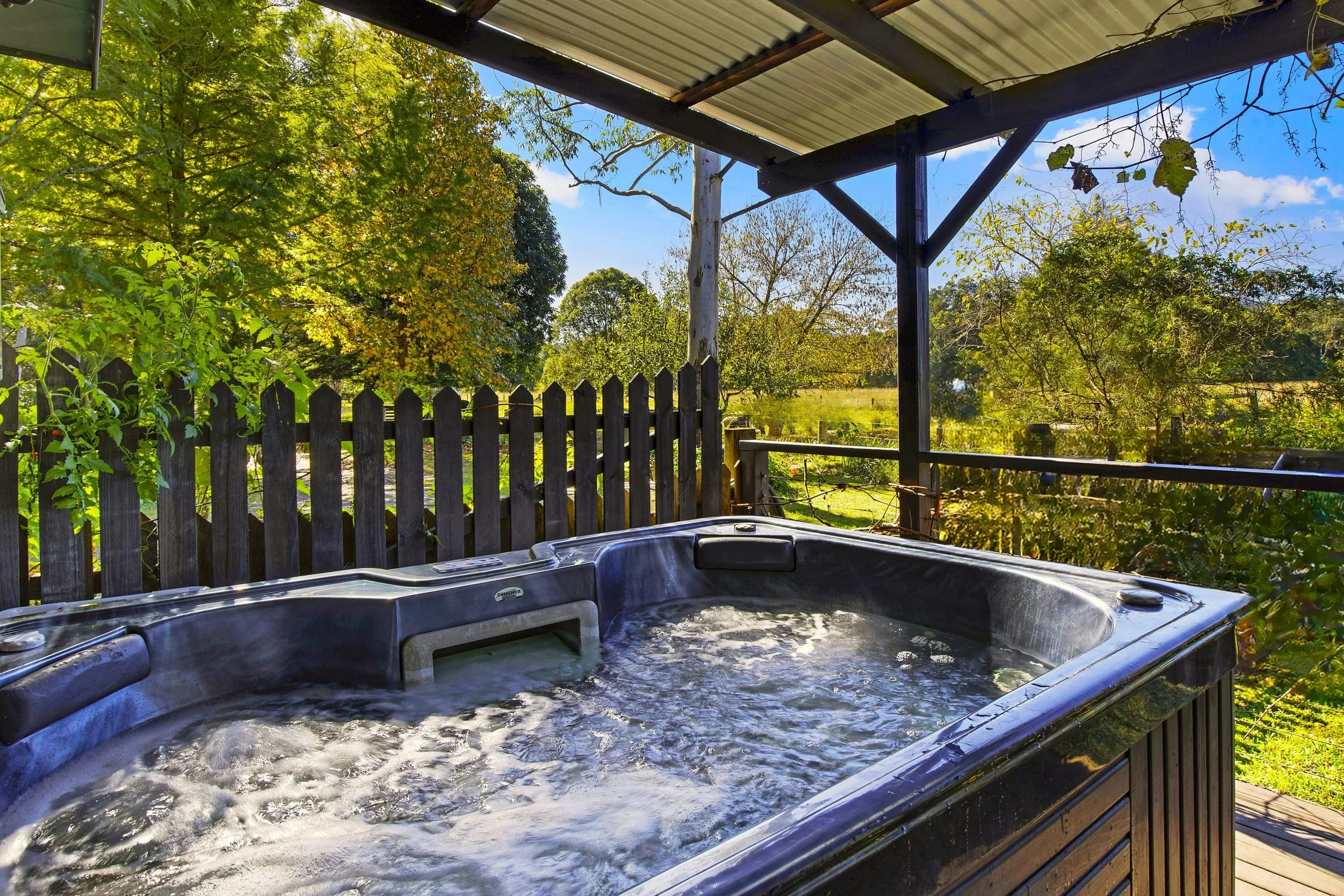 Outdoor hot tub on a deck overlooking a lush green landscape with trees and a wooden fence, under a metal roof.