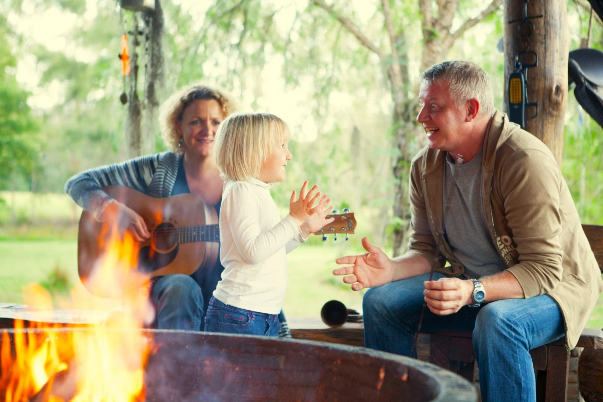 A family outdoors near a campfire, with a woman playing a guitar, a young girl talking to a man, and trees in the background.
