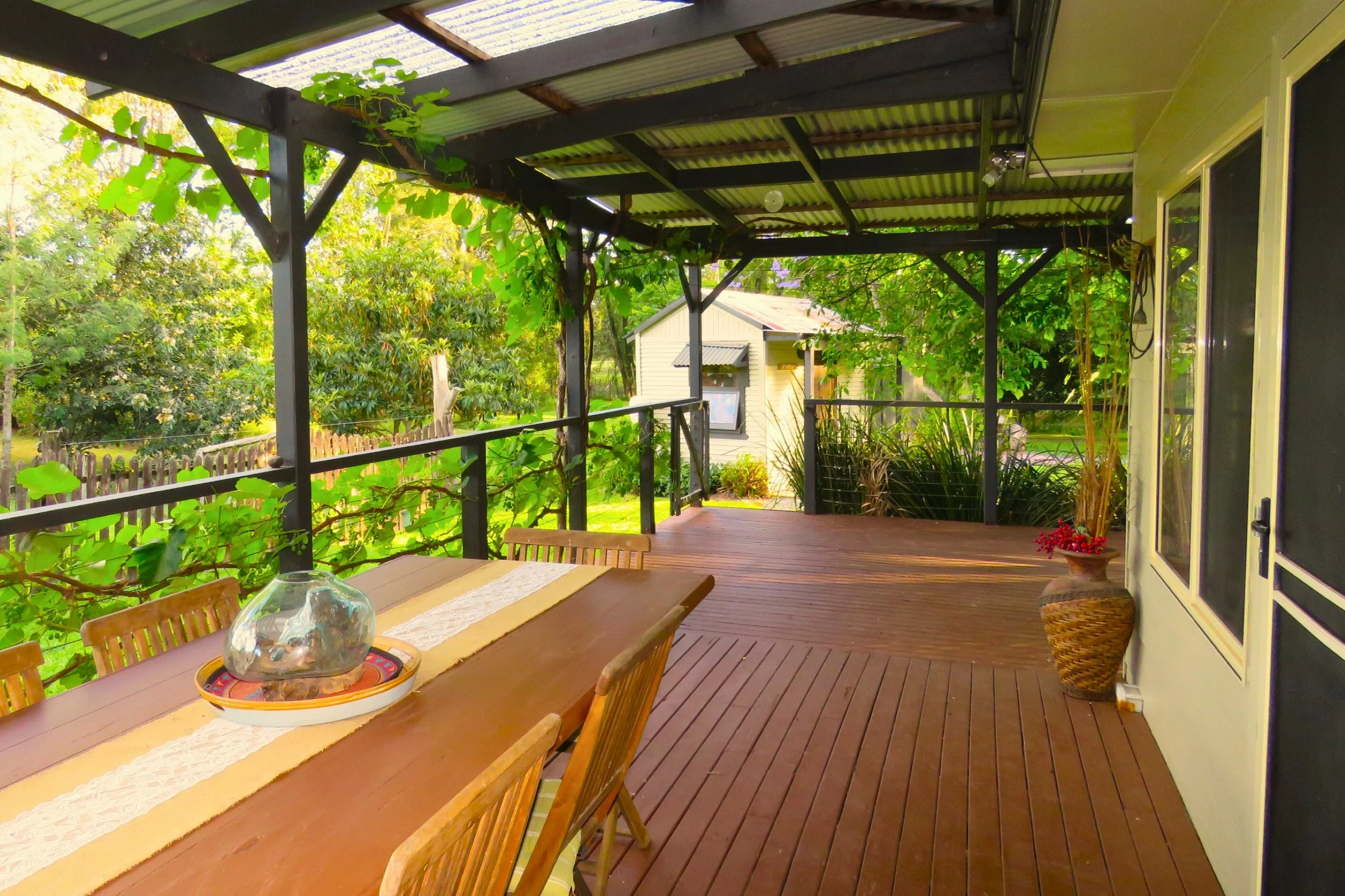 A spacious wooden porch with a dining table and chairs, surrounded by lush green trees and plants, featuring a vine-covered railing and a decorative vase with flowers.