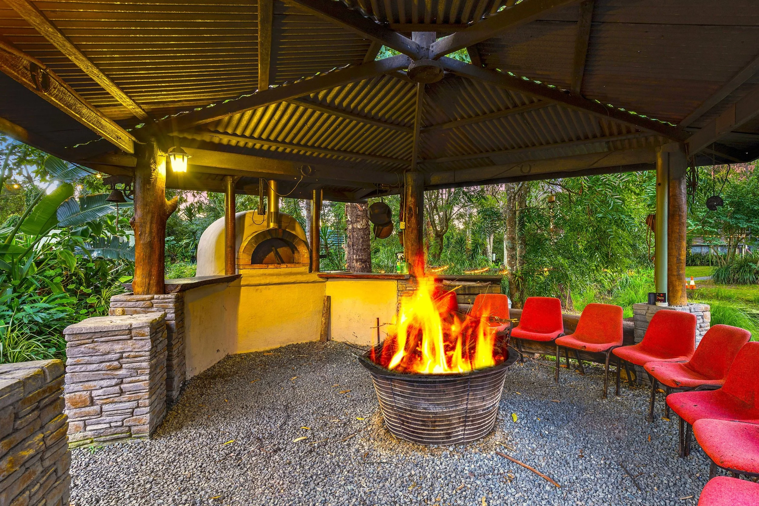 Outdoor seating area with a fire pit, red chairs, a wood-fired pizza oven, and lush greenery surrounding the space.