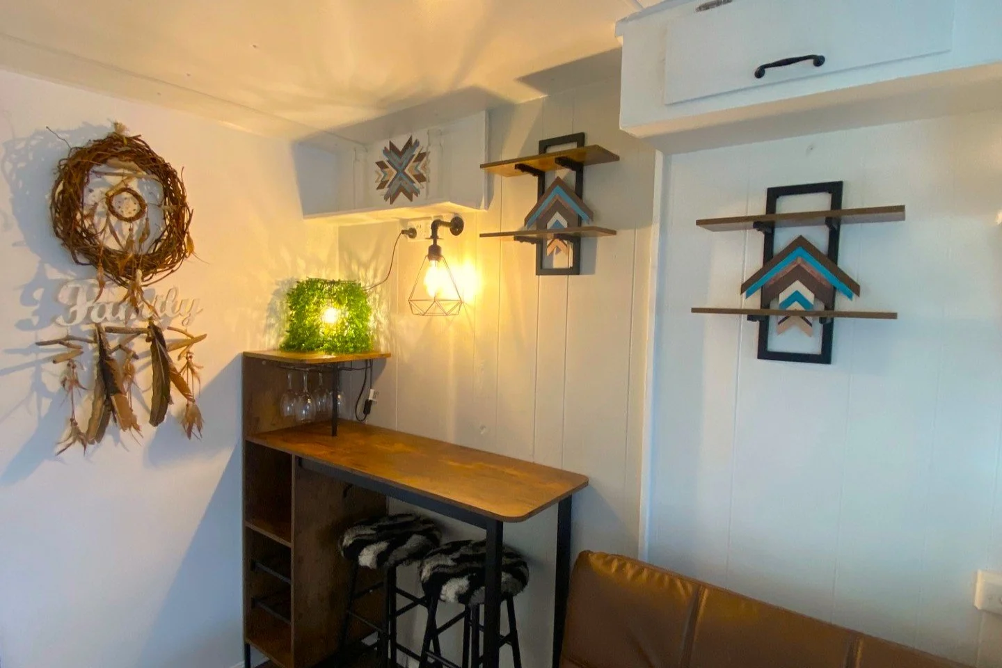 Cozy corner with a small wooden bar table, two stools with cowhide cushions, wall decorations including a wreath, a "Beautiful" sign, and various geometric wall shelves with framed arrow artwork, illuminated by a hanging vintage-style lightbulb.