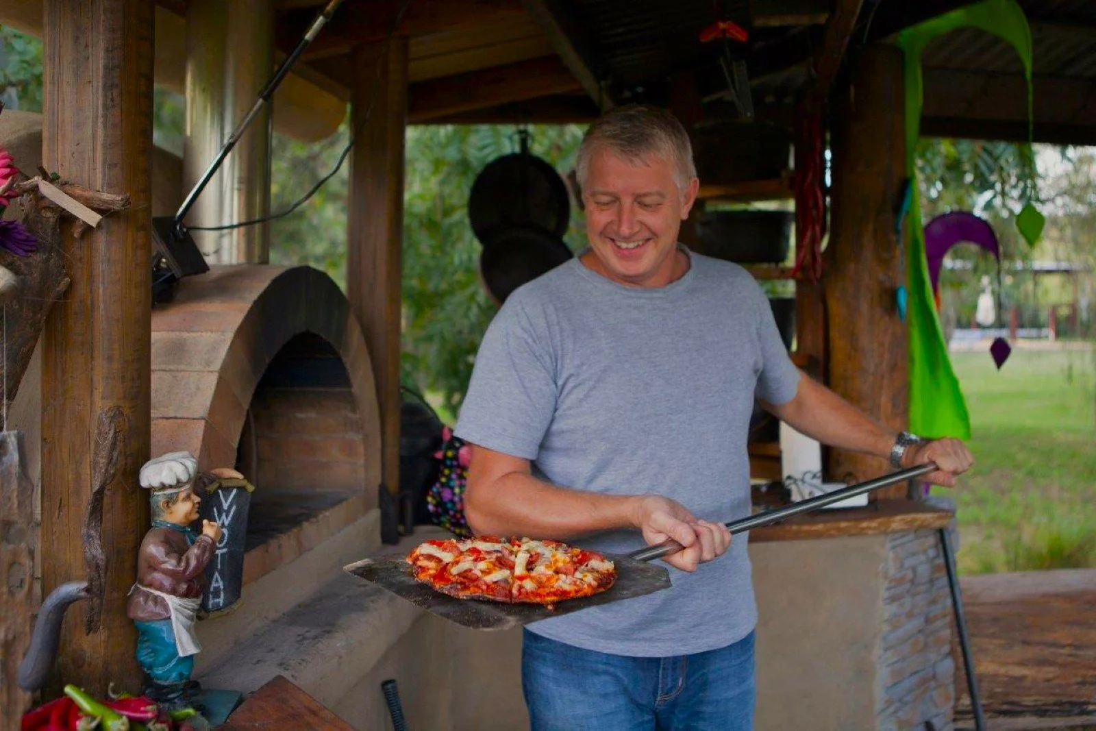 A man with gray hair wearing a gray t-shirt baking pizza on a pizza peel at an outdoor stone and wood pizza oven, smiling and looking down.