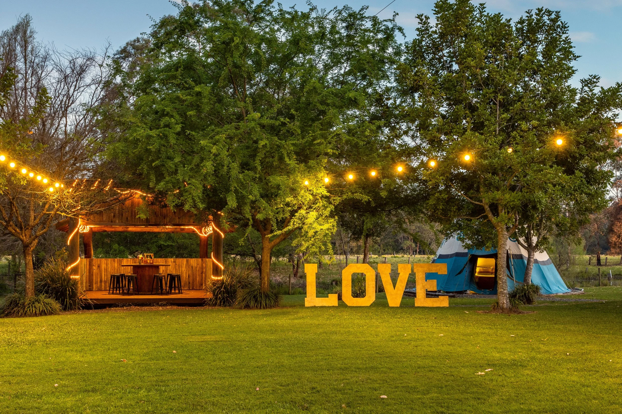 Outdoor scene with string lights hanging over a wooden bar area, large yellow 'LOVE' letters on the grass, a blue and white tent, and trees in the evening.