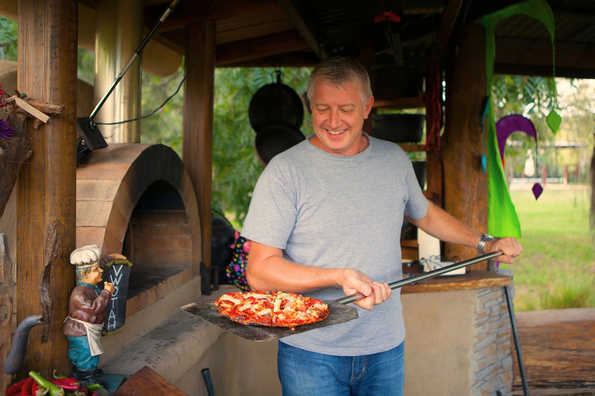 A man in a gray t-shirt smiling while holding a pizza peel with a freshly baked pizza at an outdoor pizza oven.