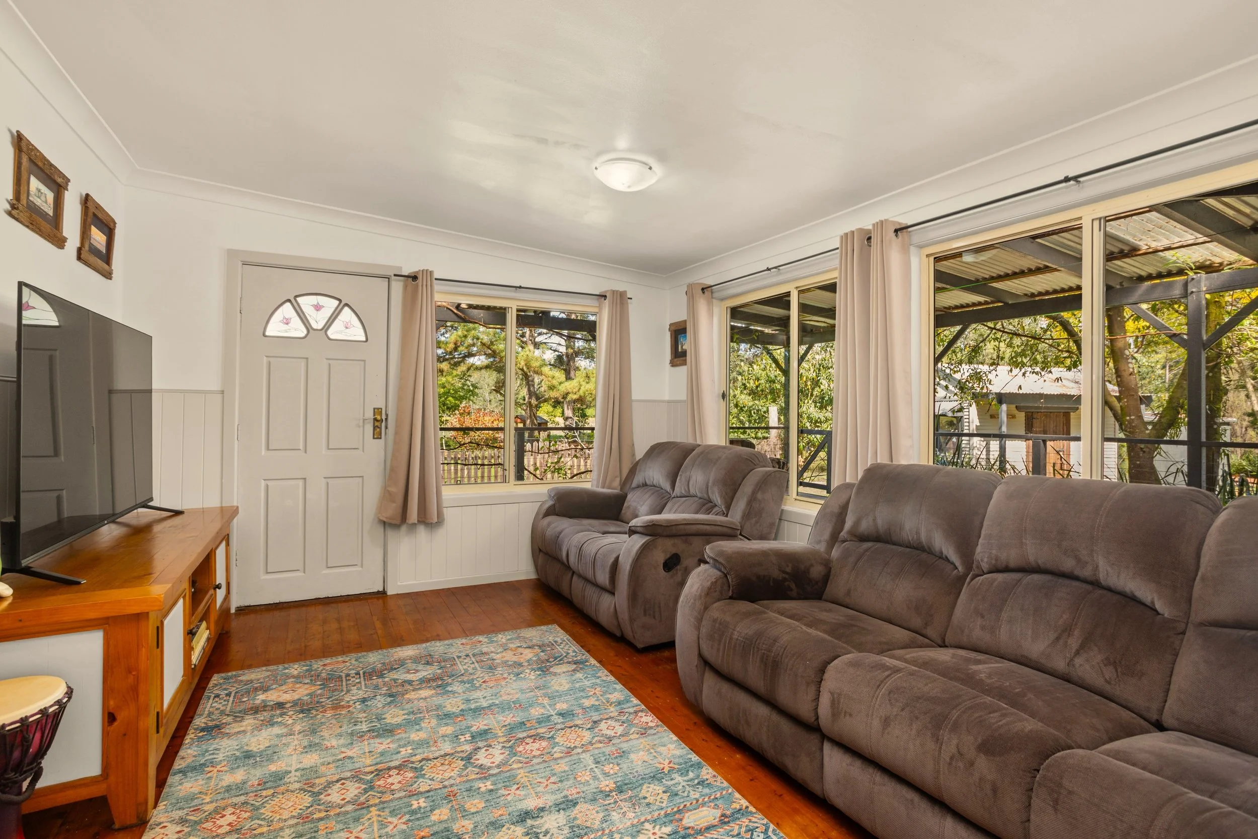 Living room with three brown sofas, large windows with beige curtains, a wooden TV stand with a flat-screen TV, and a colorful area rug on hardwood floor, with trees visible outside.