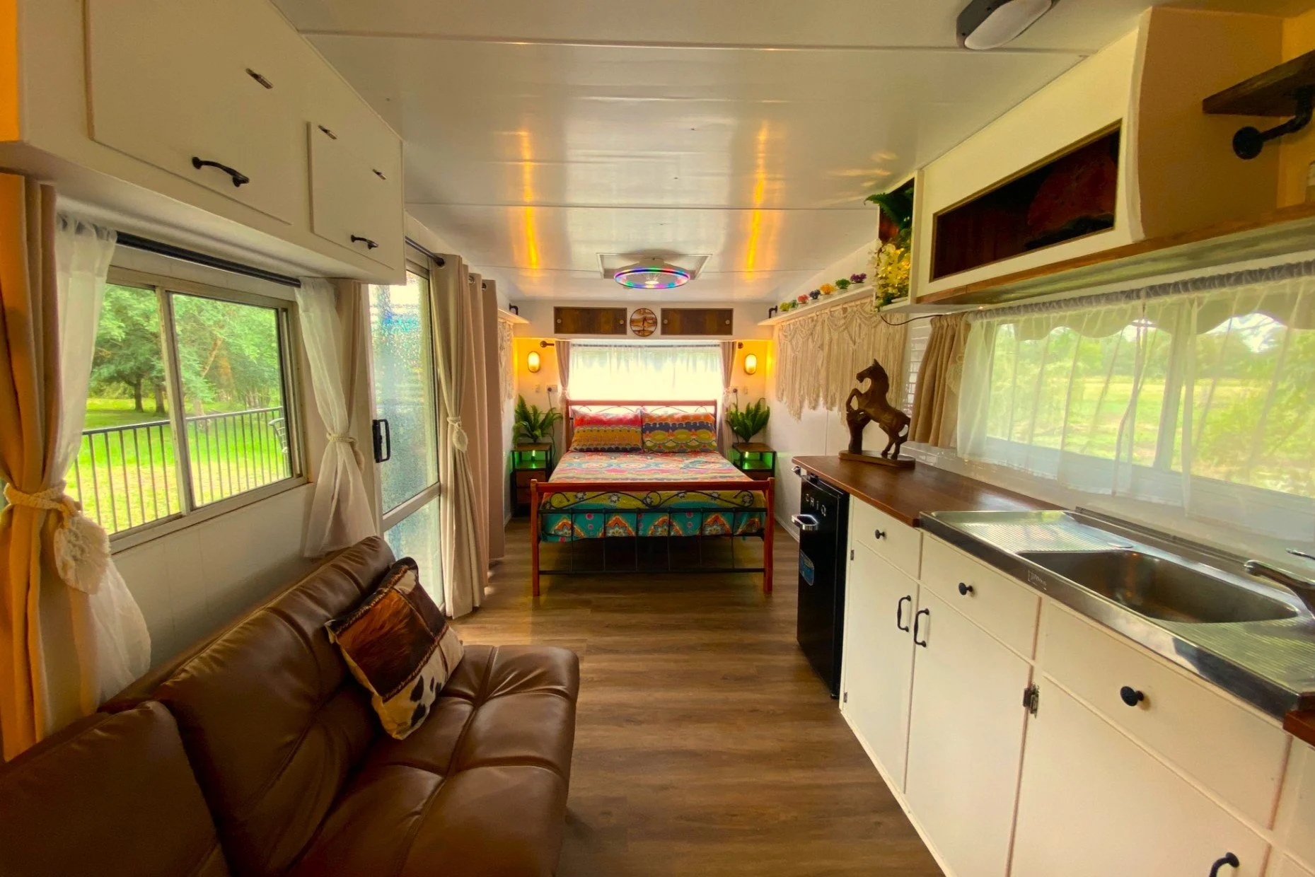 Interior of a cozy cabin or tiny home with a sleeping area at the back, a kitchen area on the right, and a small living space with a brown leather sofa on the left. The room is decorated with plants, a horse sculpture, colorful bedding, and natural l