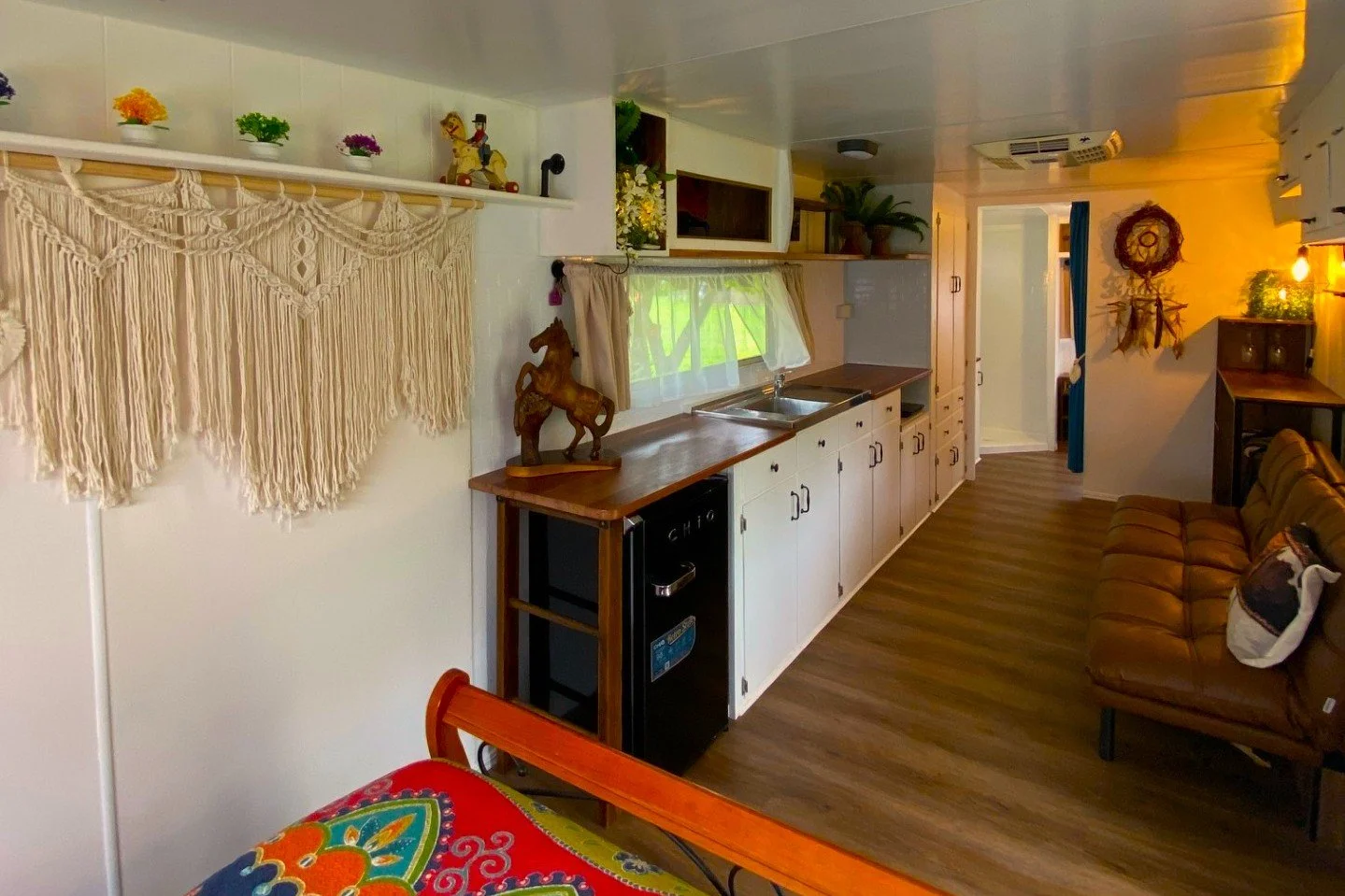 Cozy kitchen and living space with wooden cabinets, a small refrigerator, a brown leather couch, wall decorations, and a window with curtains