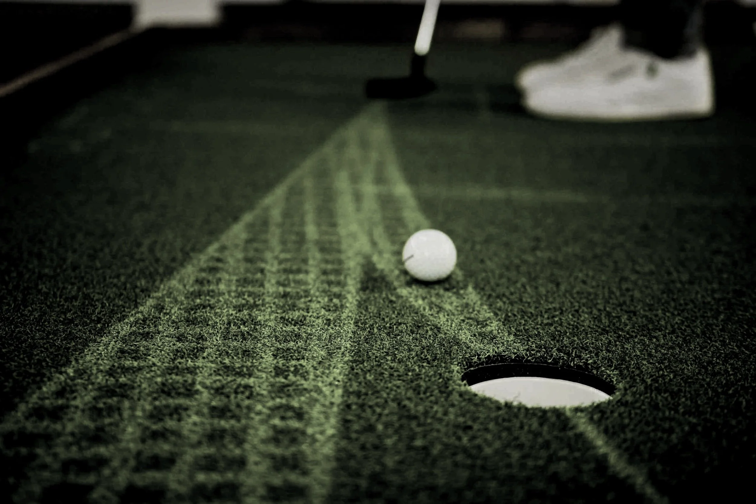 Close-up of a golf ball on an artificial turf mat near a hole, with a putter and a person wearing white sneakers in the background.