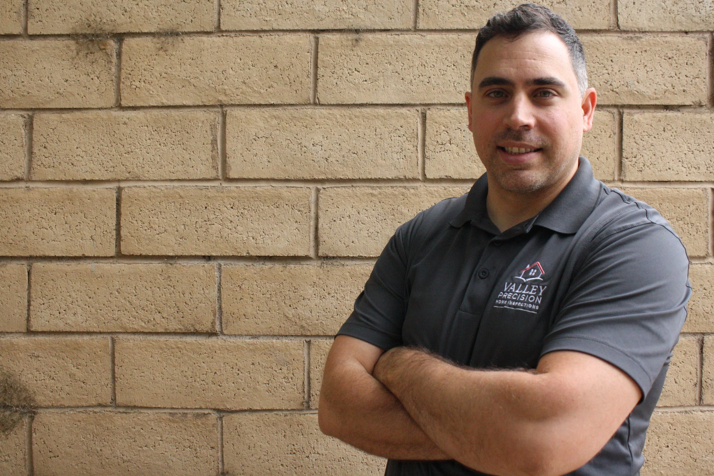 A man with short dark hair smiling and crossing his arms in front of a beige brick wall. He is wearing a dark polo shirt with a logo reading 'Valley Precision Home Inspections'.