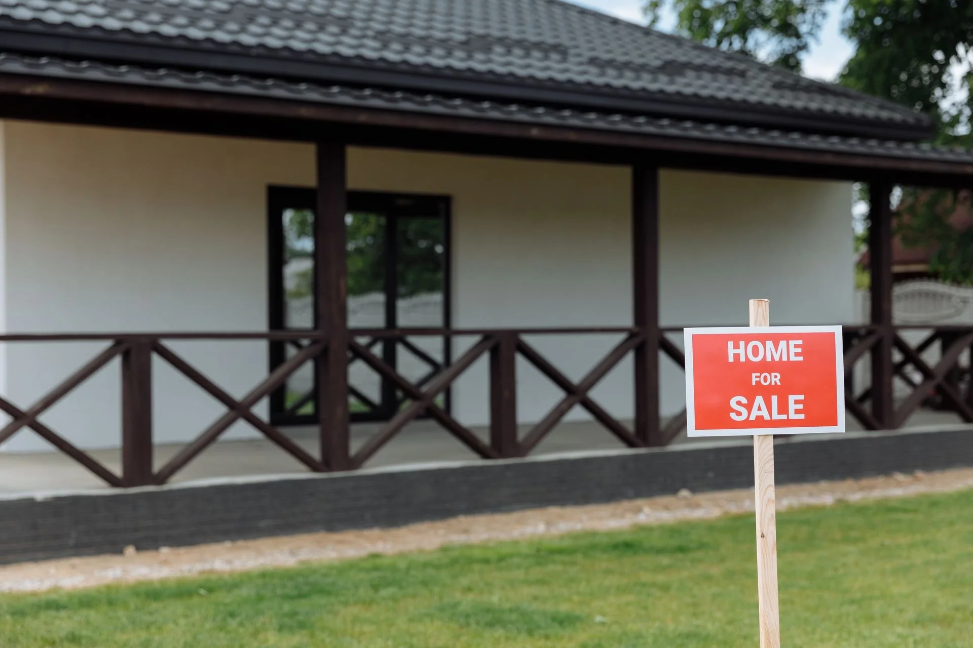 A house with a dark roof, beige walls, and large windows, with a "Home for Sale" sign in the front yard.