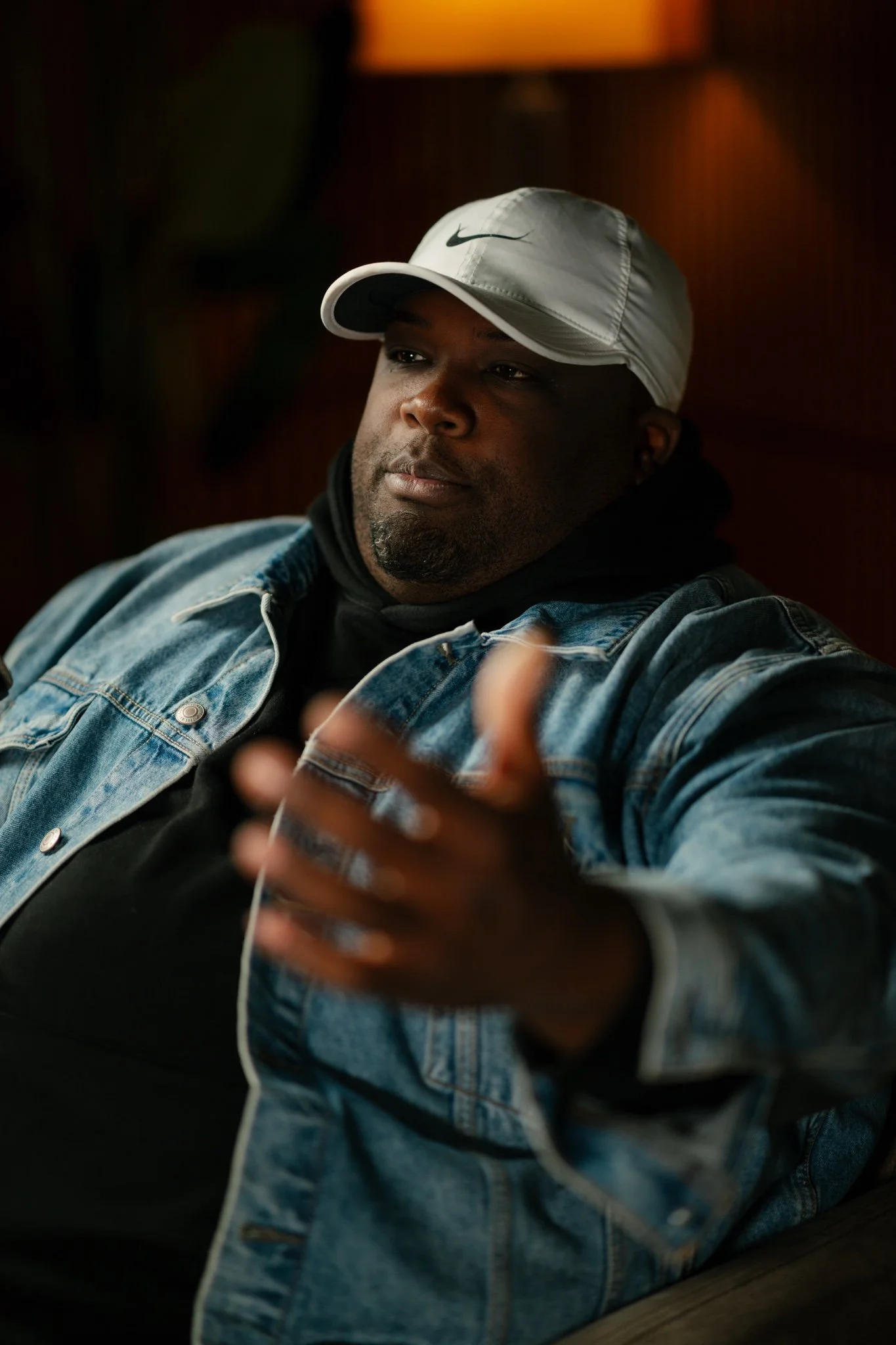 A man wearing a white Nike cap, black hoodie, and denim jacket sitting indoors pointing towards the camera with a confident expression.