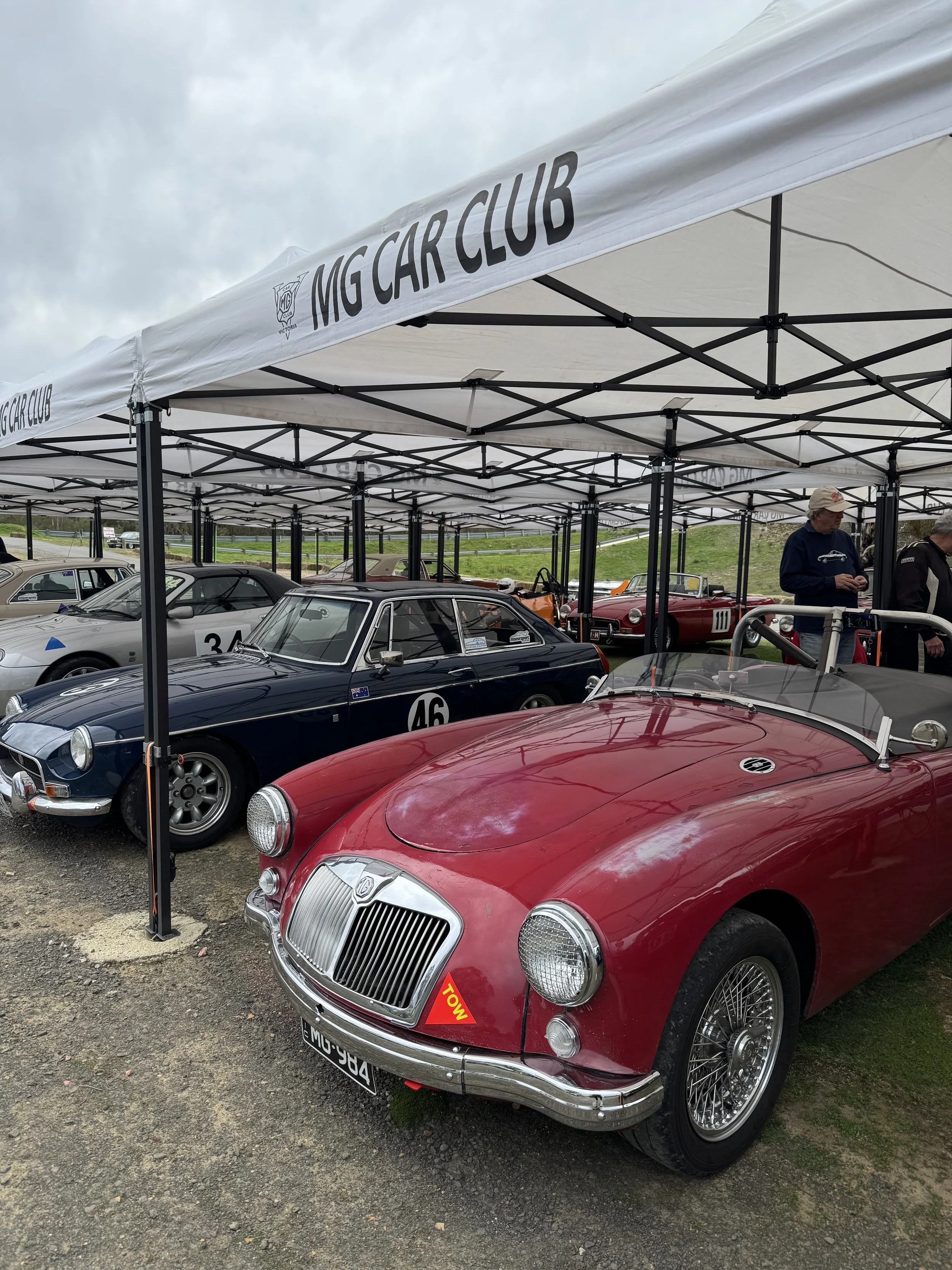 Vintage red convertible car parked at MG Car Club event with other classic cars and spectators under a white tent