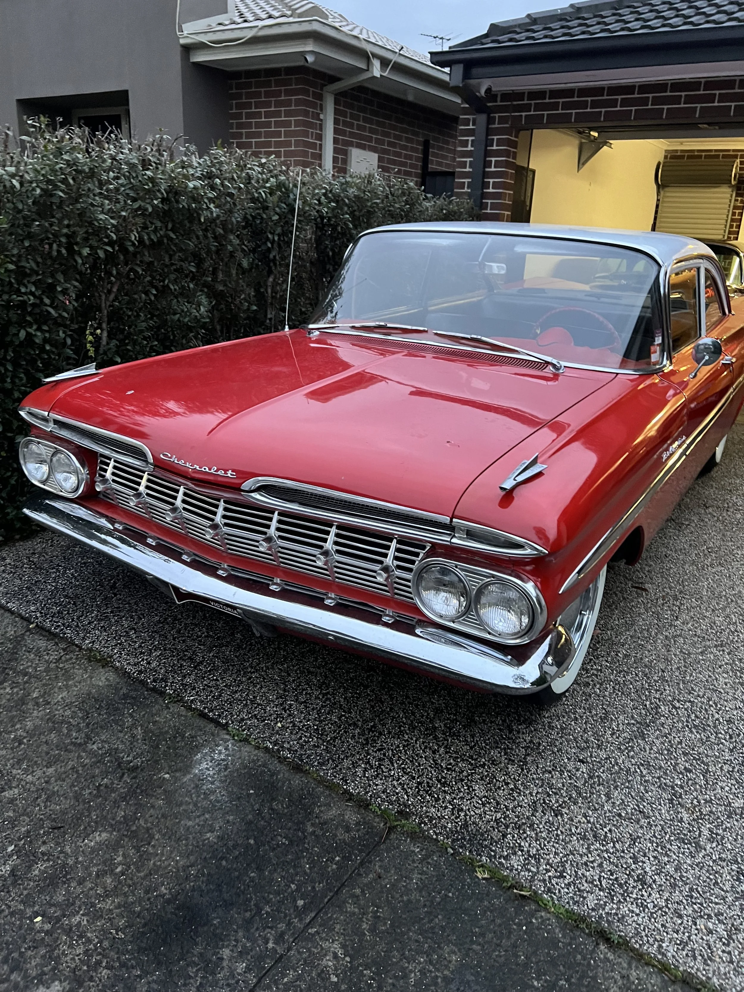 A vintage red Chevrolet car parked on a driveway next to a house, with a hedge on the left and an open garage on the right.