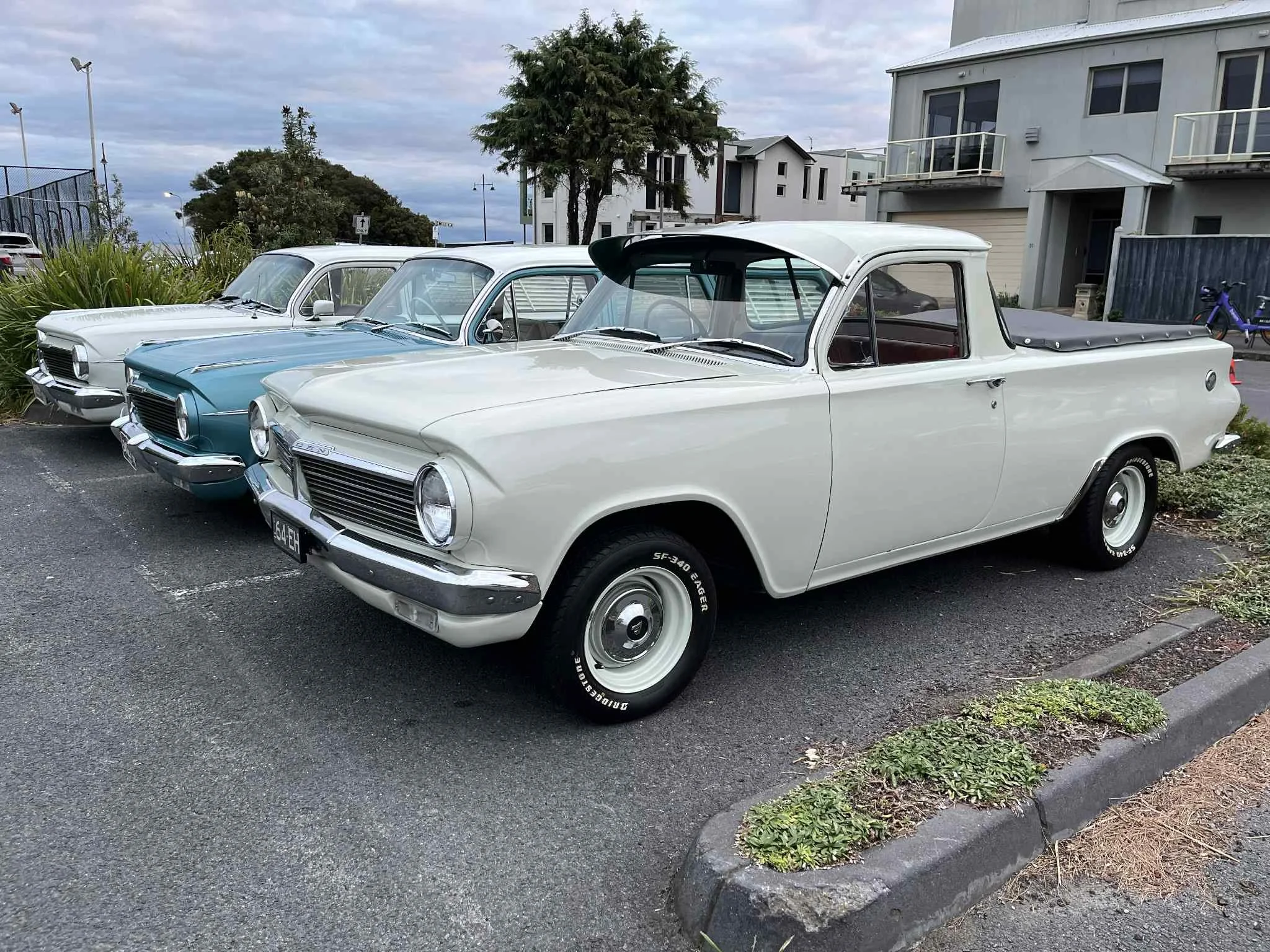 Three vintage cars parked in a row next to a sidewalk with residential buildings in the background, featuring a white pickup truck in the foreground and two other classic cars behind it.