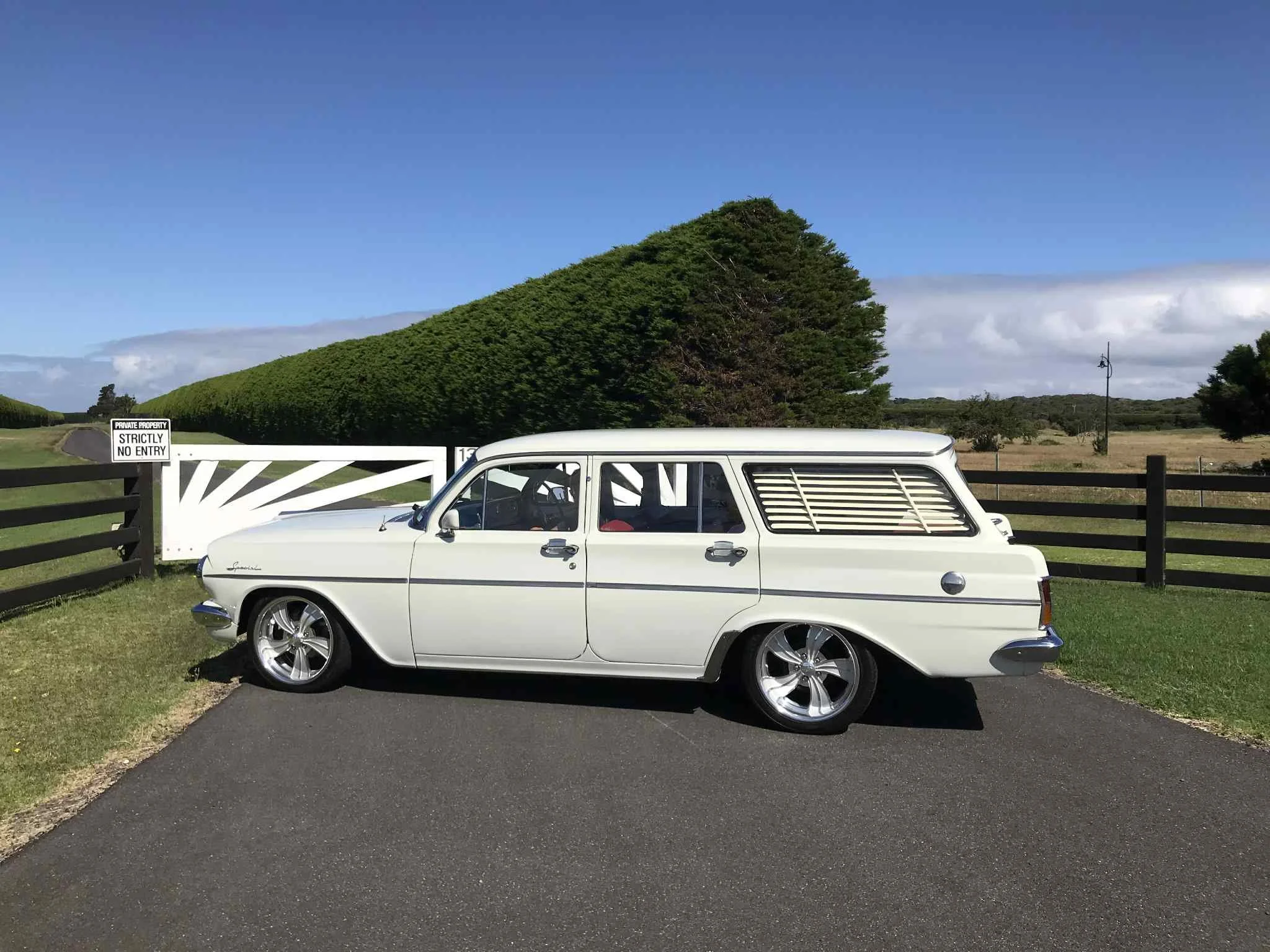 A vintage white station wagon parked on a paved surface in front of a wooden fence, with a hedge and open field in the background under a partly cloudy sky.