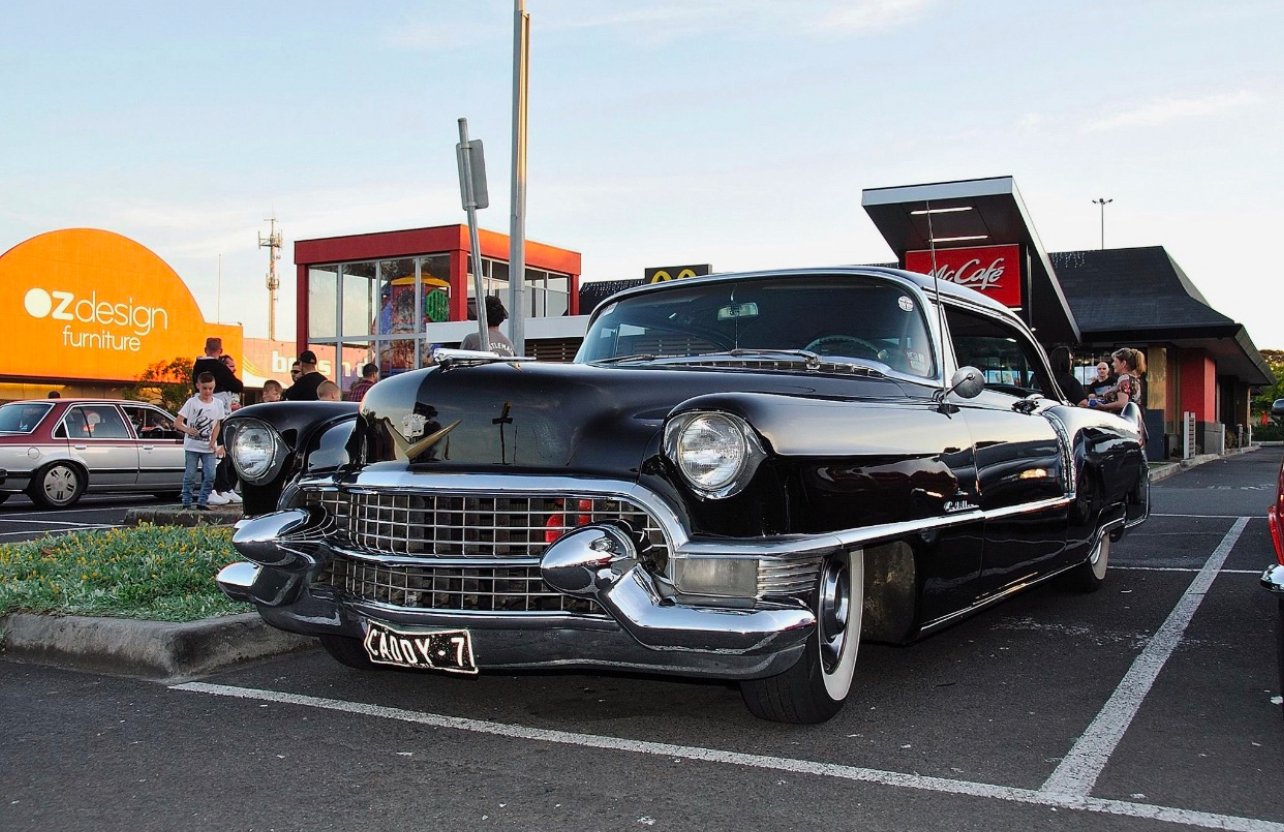 A vintage black Cadillac parked in a parking lot during daytime with people in the background near a McDonald's and oz design furniture store.