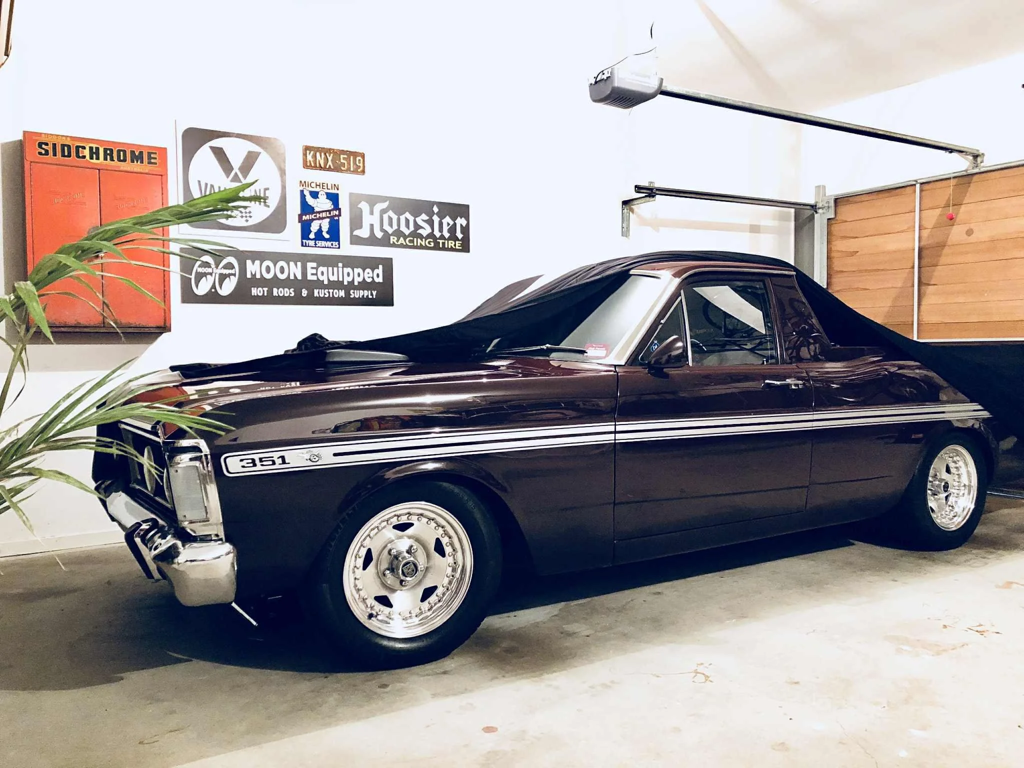 A black vintage car with a roof cover parked in a garage, with automotive and racing signs on the wall behind it.