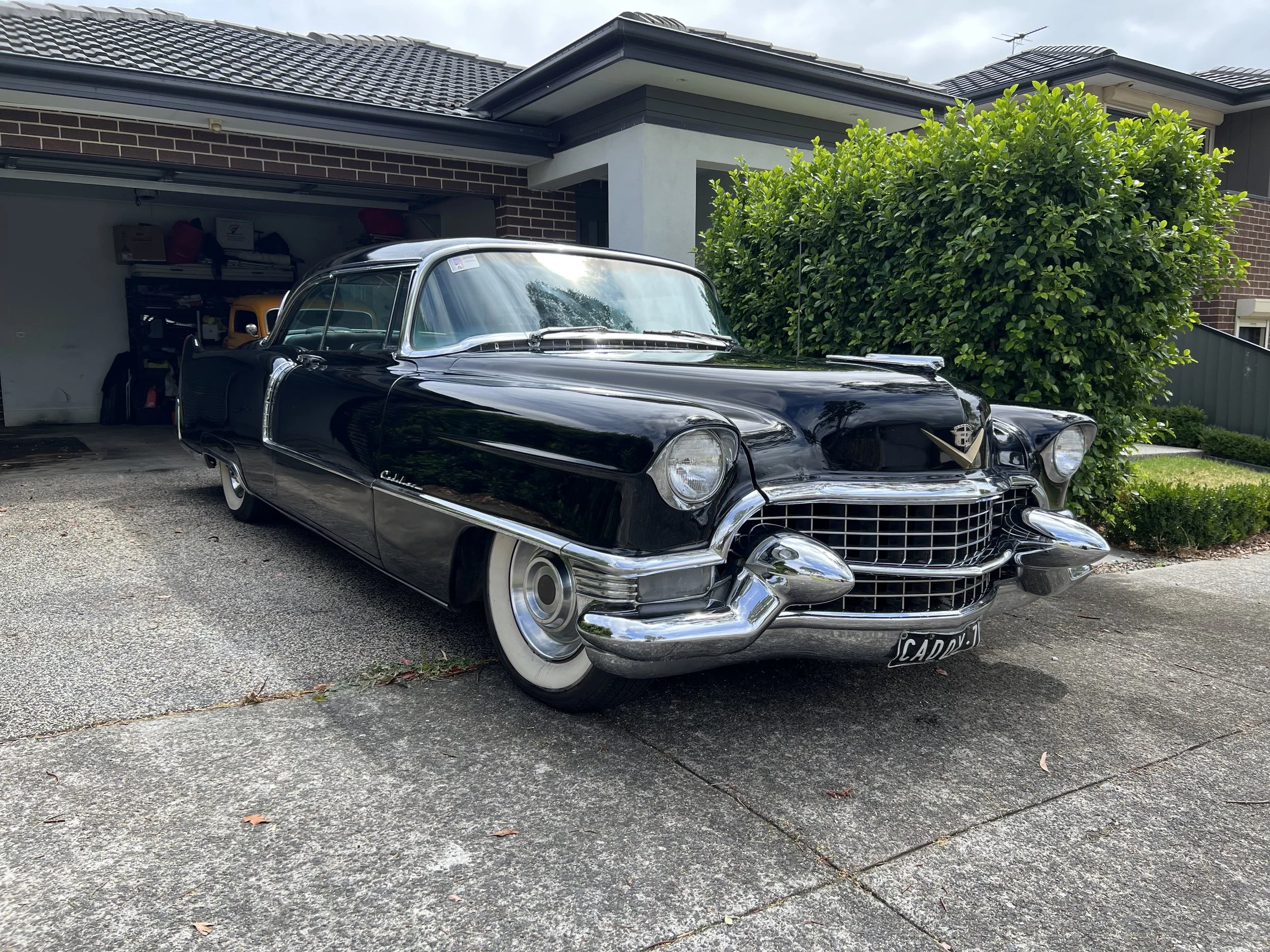 A vintage black Cadillac parked in a driveway in front of a house with a garage. The car has chrome accents and white-wall tires, with a garden bush to its right.