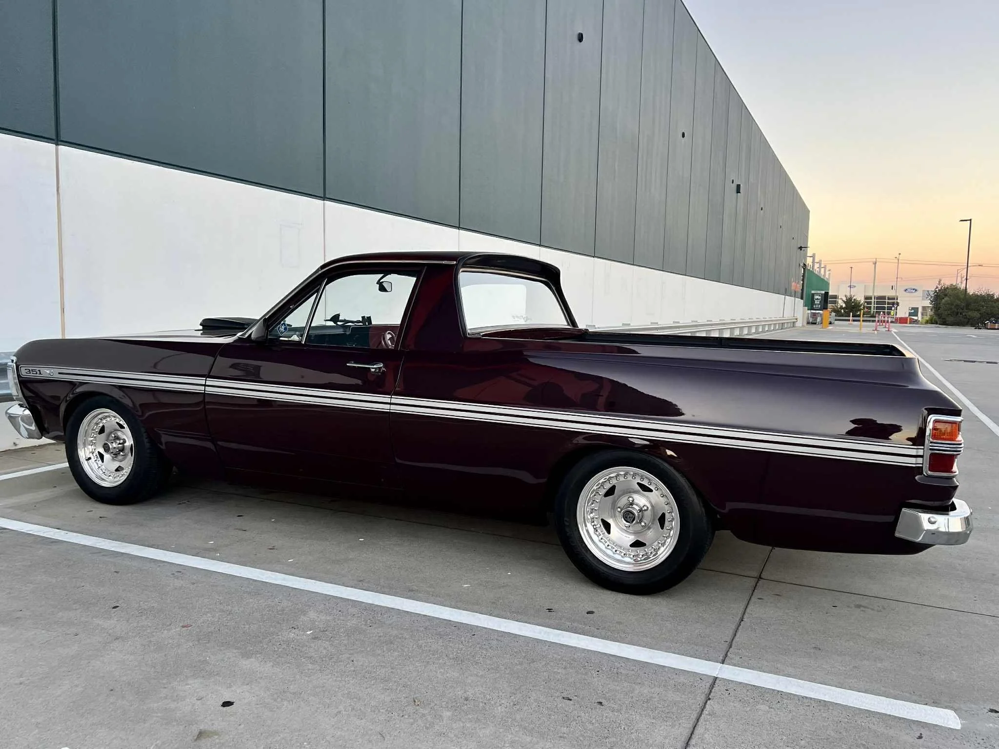 A vintage dark red pickup truck parked in a parking lot near a modern building during sunset.