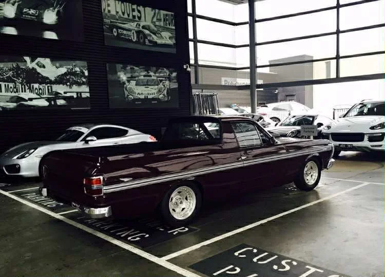 A vintage maroon car parked inside a modern showroom with several other luxury cars, large glass windows letting in natural light, and vintage racing posters on the wall.