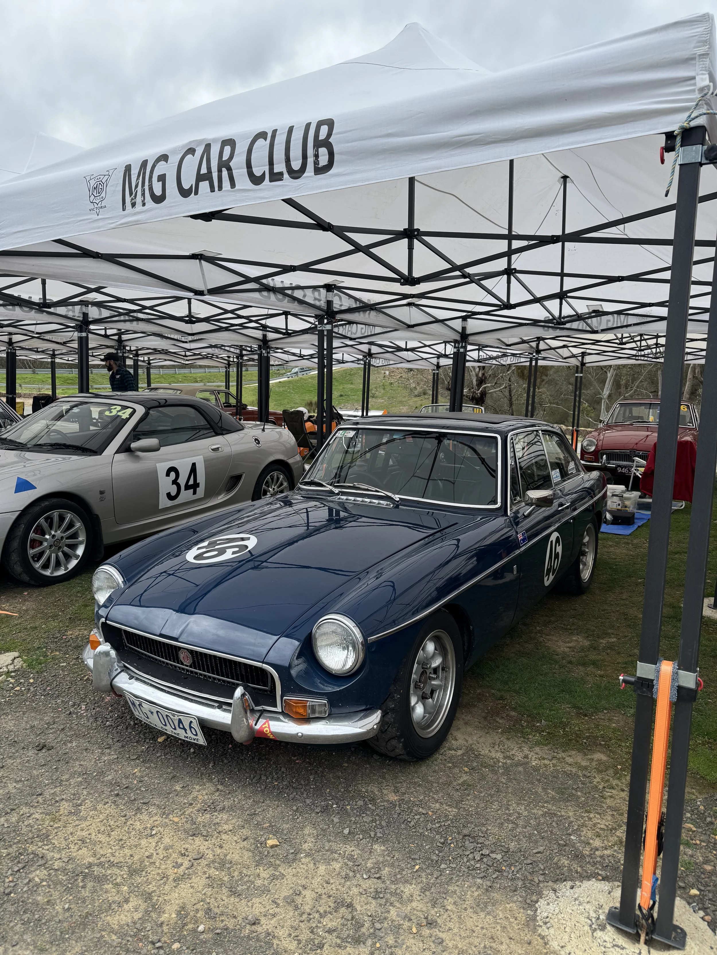 Vintage blue MG car with racing number 46 at an outdoor car show under a white tent with MG Car Club banner, alongside other classic cars.