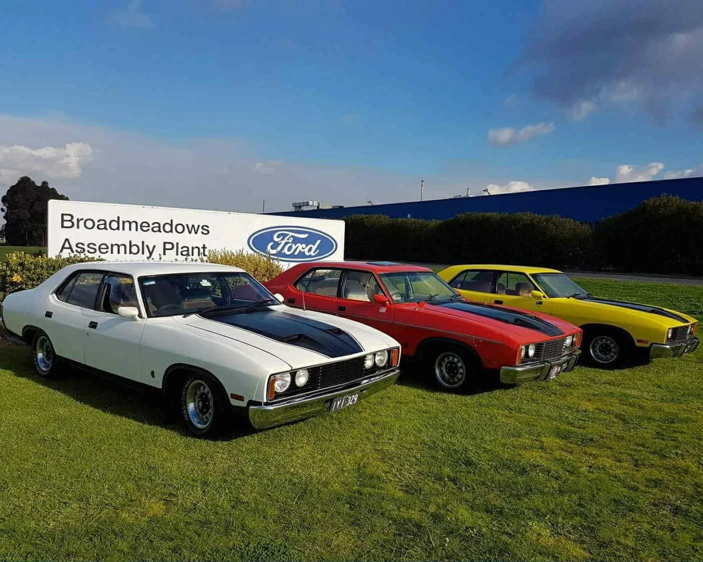 Three vintage cars parked on grass in front of a Ford sign that reads Broadmeadows Assembly Plant, with a blue building and cloudy sky in the background.
