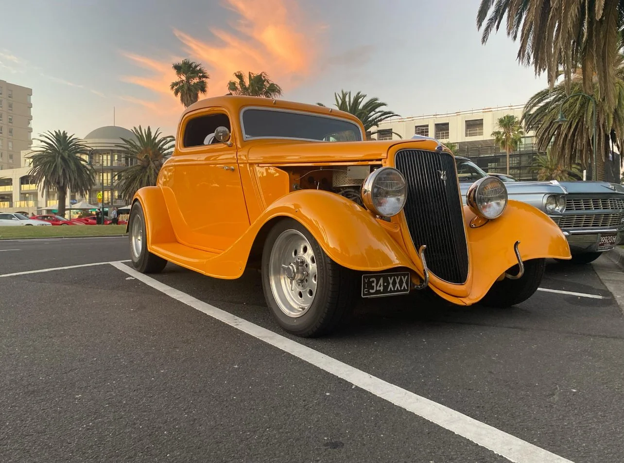 Orange vintage car parked in a parking lot during sunset, with palm trees and buildings in the background.