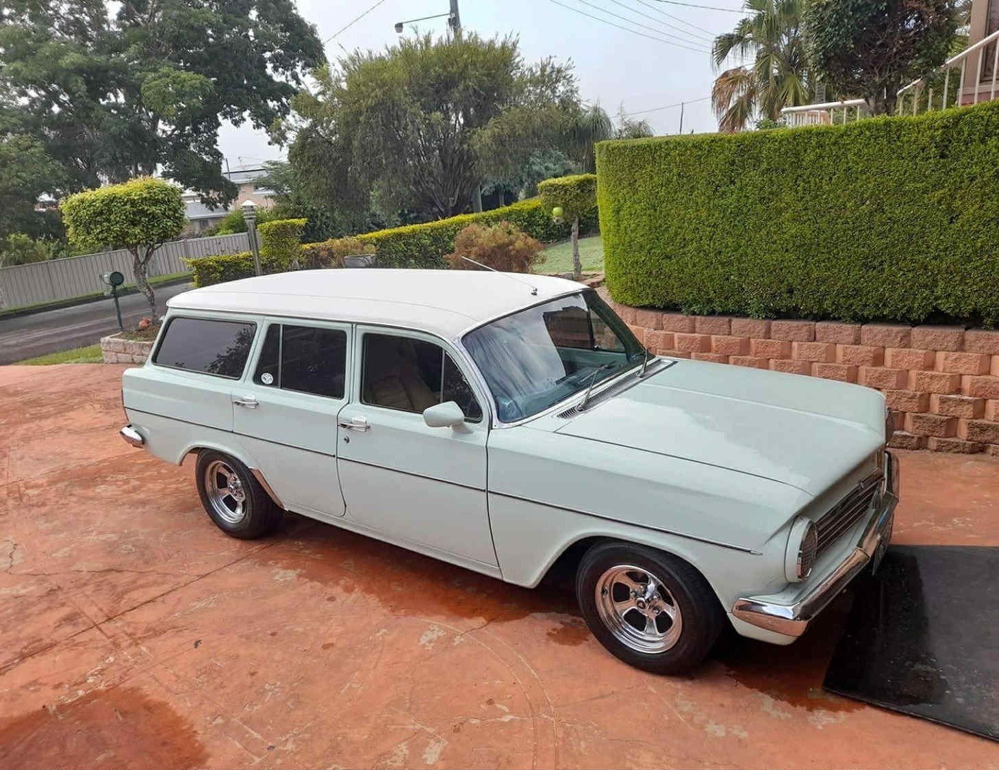 White vintage station wagon parked on a driveway with green bushes and trees in the background.