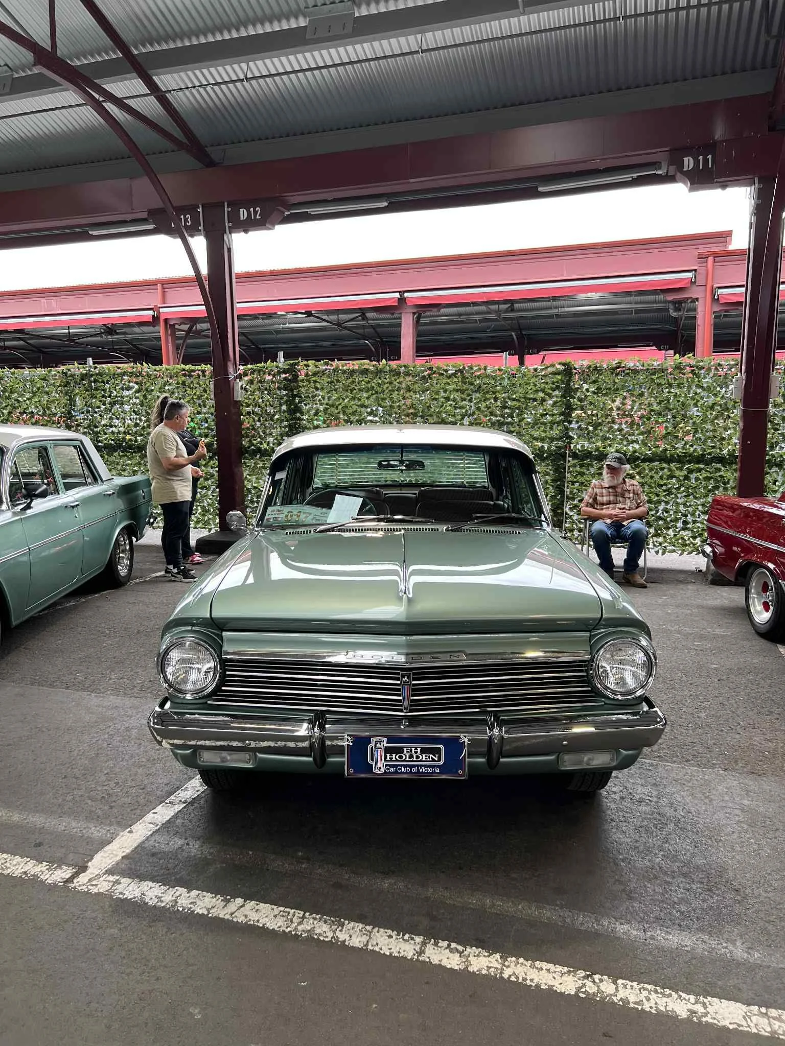 A vintage green Holden car on display at a car show, with a license plate reading 'HOLDEN' and a sign in the windshield, situated in an indoor parking area.