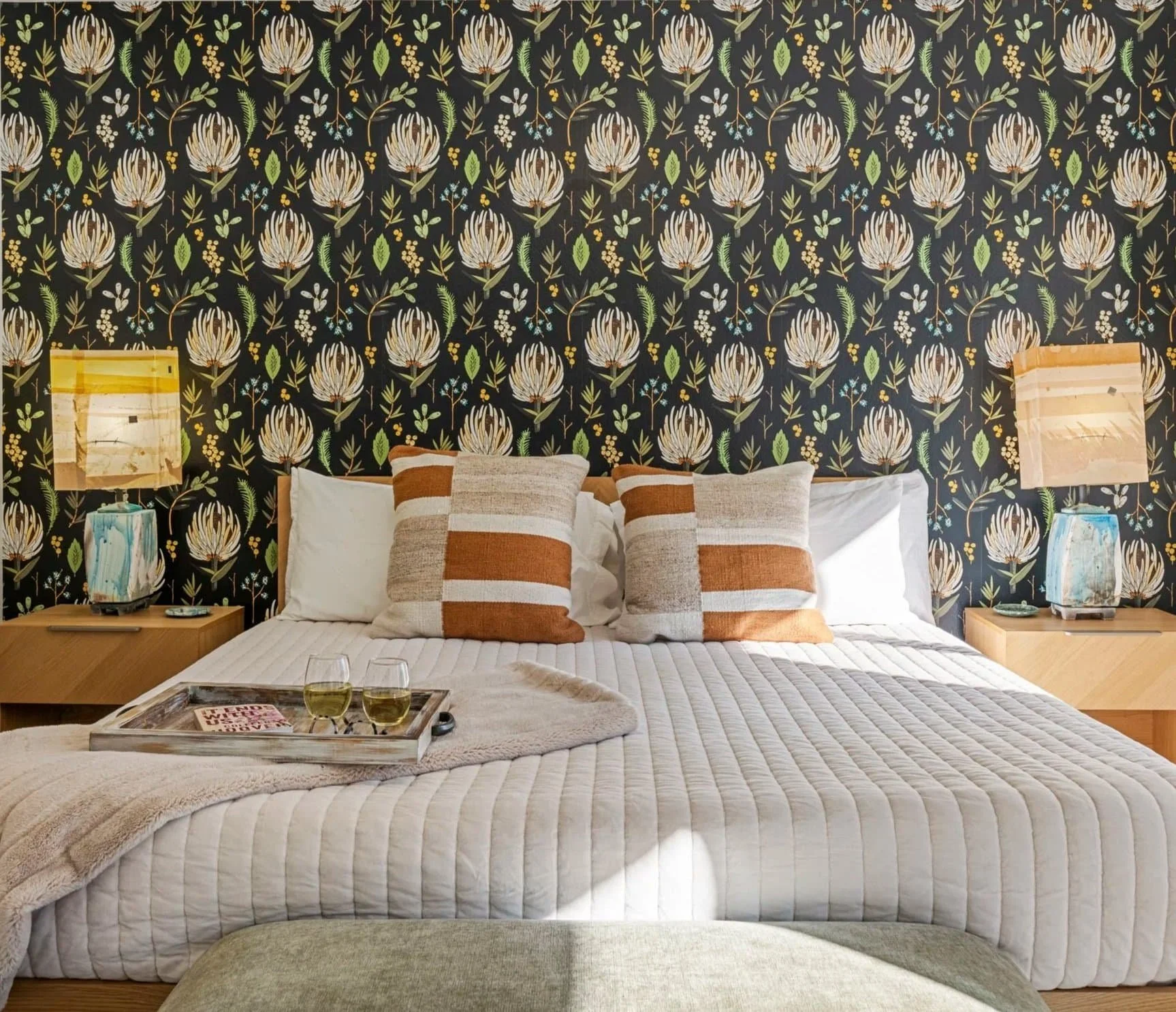 A neatly made bed with two striped pillows, a tray with two glasses of white wine, a book, and eyeglasses, a wooden nightstand with a colorful lamp and decorative item, against a black floral wallpaper background.