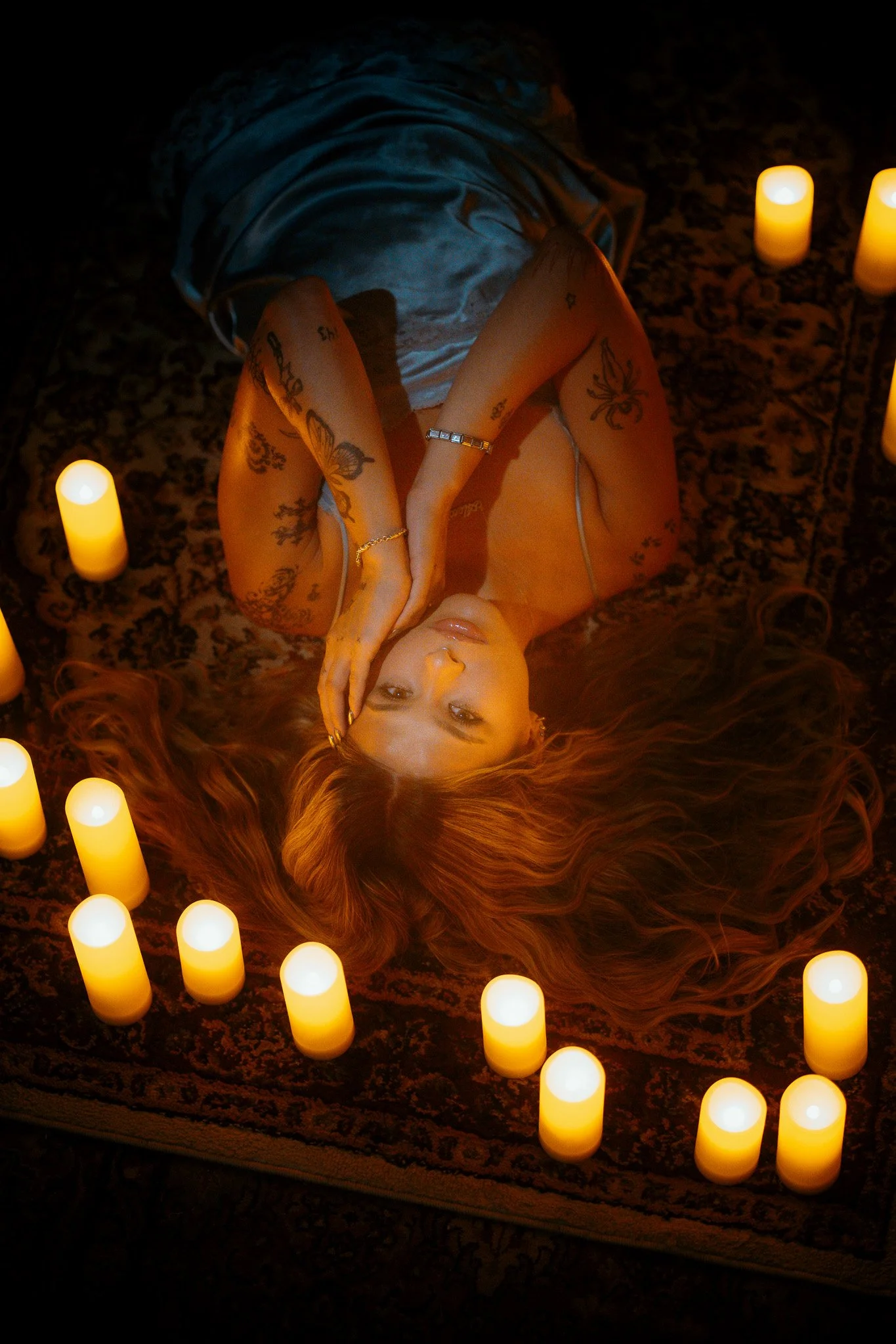 A woman with long red hair lying on her back on a patterned rug, surrounded by candles, covering one eye with her hand, tattoos on her arms, and wearing a jewelry bracelet and a light-colored garment.