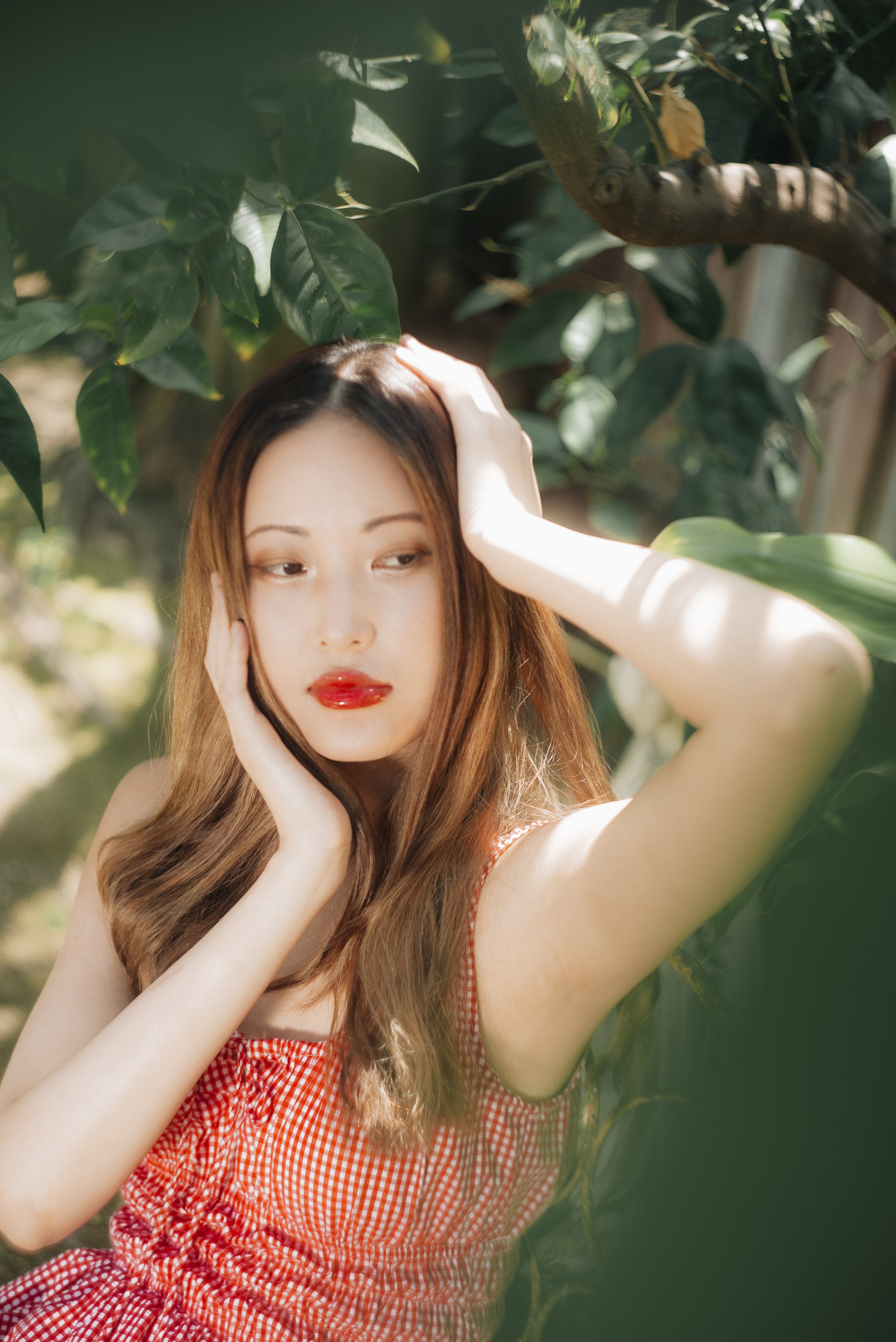 A young woman with long, wavy brown hair and red lipstick, wearing a red and white gingham dress, standing in a lush green outdoor setting with leaves and sunlight, gently touching her head and face.