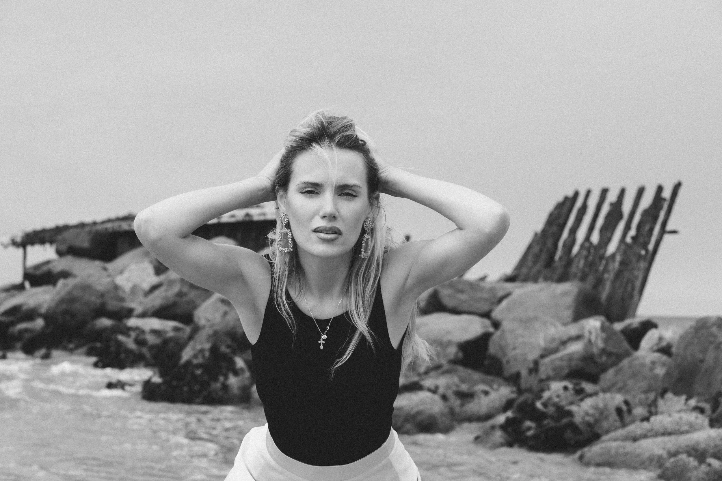 Black and white photo of a woman with long hair, wearing large earrings, a necklace with a cross pendant, a black top, and a light-colored skirt, standing on a rocky beach with a damaged wooden structure in the background.