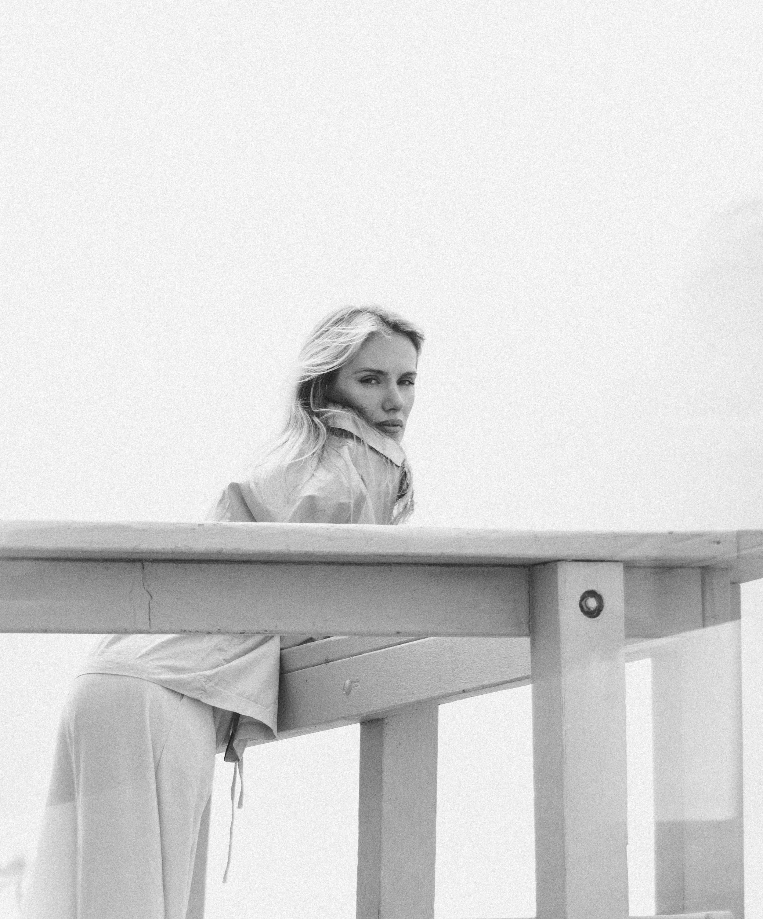 Black and white photo of a woman with wavy hair sitting on a wooden picnic table with worn paint, looking at the camera.