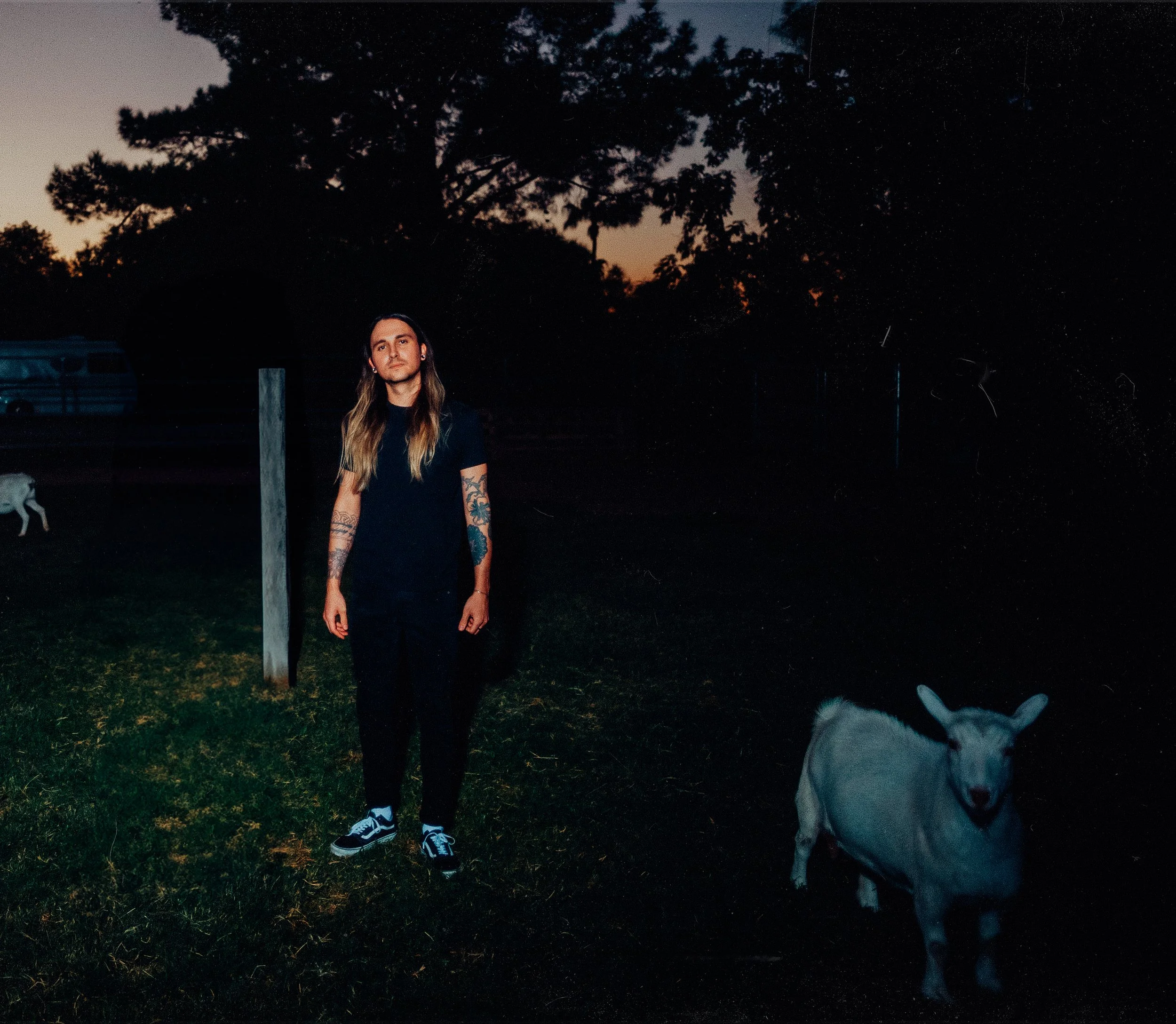 A person with long hair and tattoos on their arms standing outdoors at dusk, with trees and a multicolored sunset in the background, and a dog with a white face and darker body standing nearby.