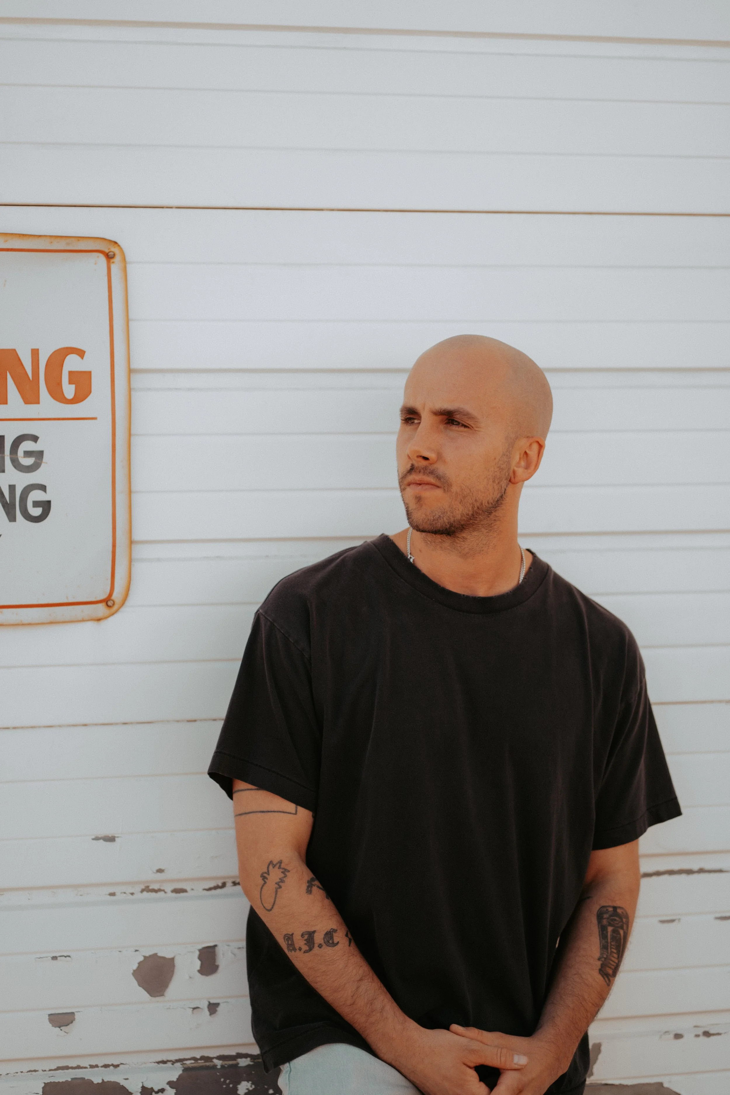 A bald man with tattoos on his arms, wearing a black t-shirt, standing in front of a white wall with peeling paint and a sign.