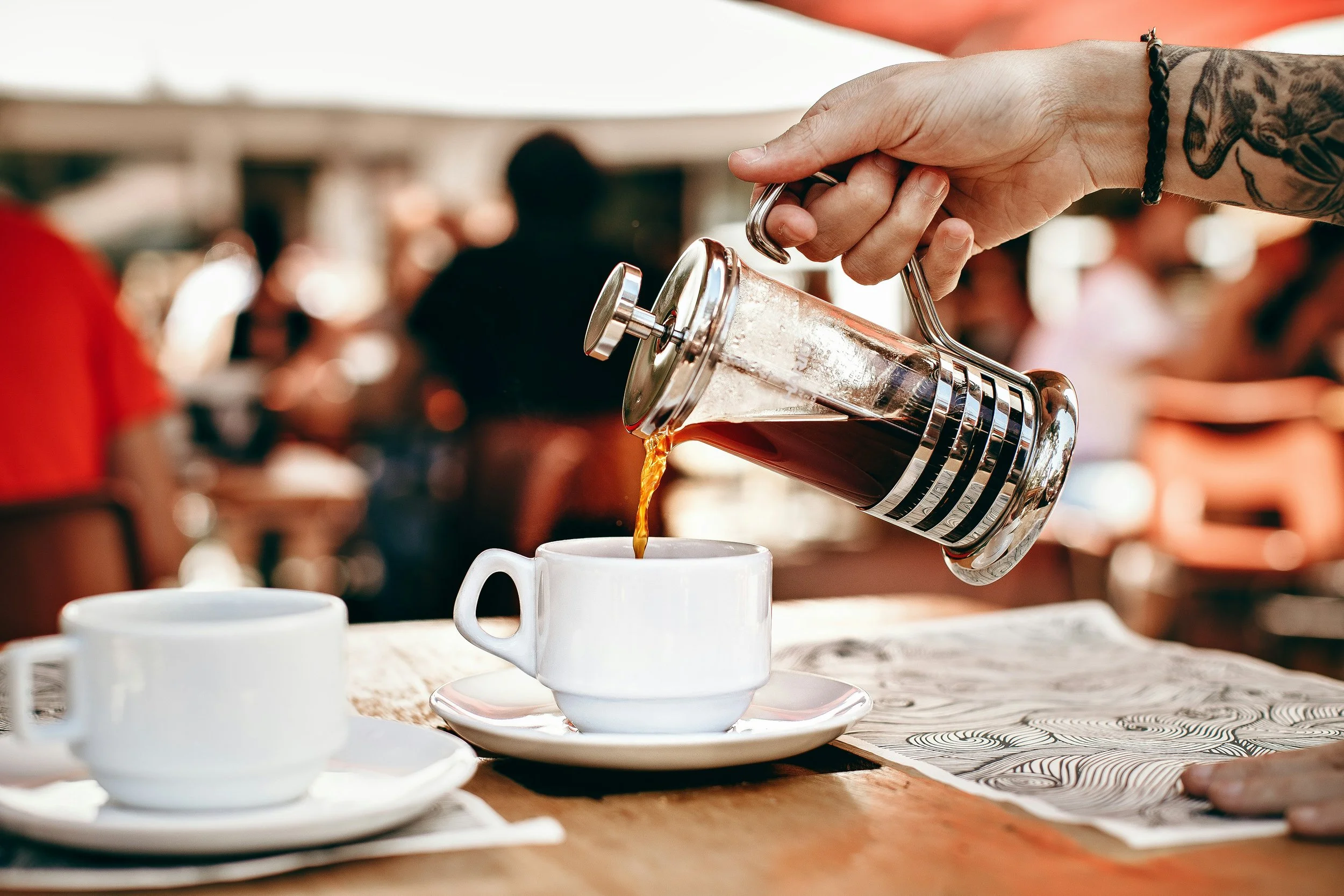 Person pouring coffee from a French press into a white coffee cup on a wooden table at a busy cafe.