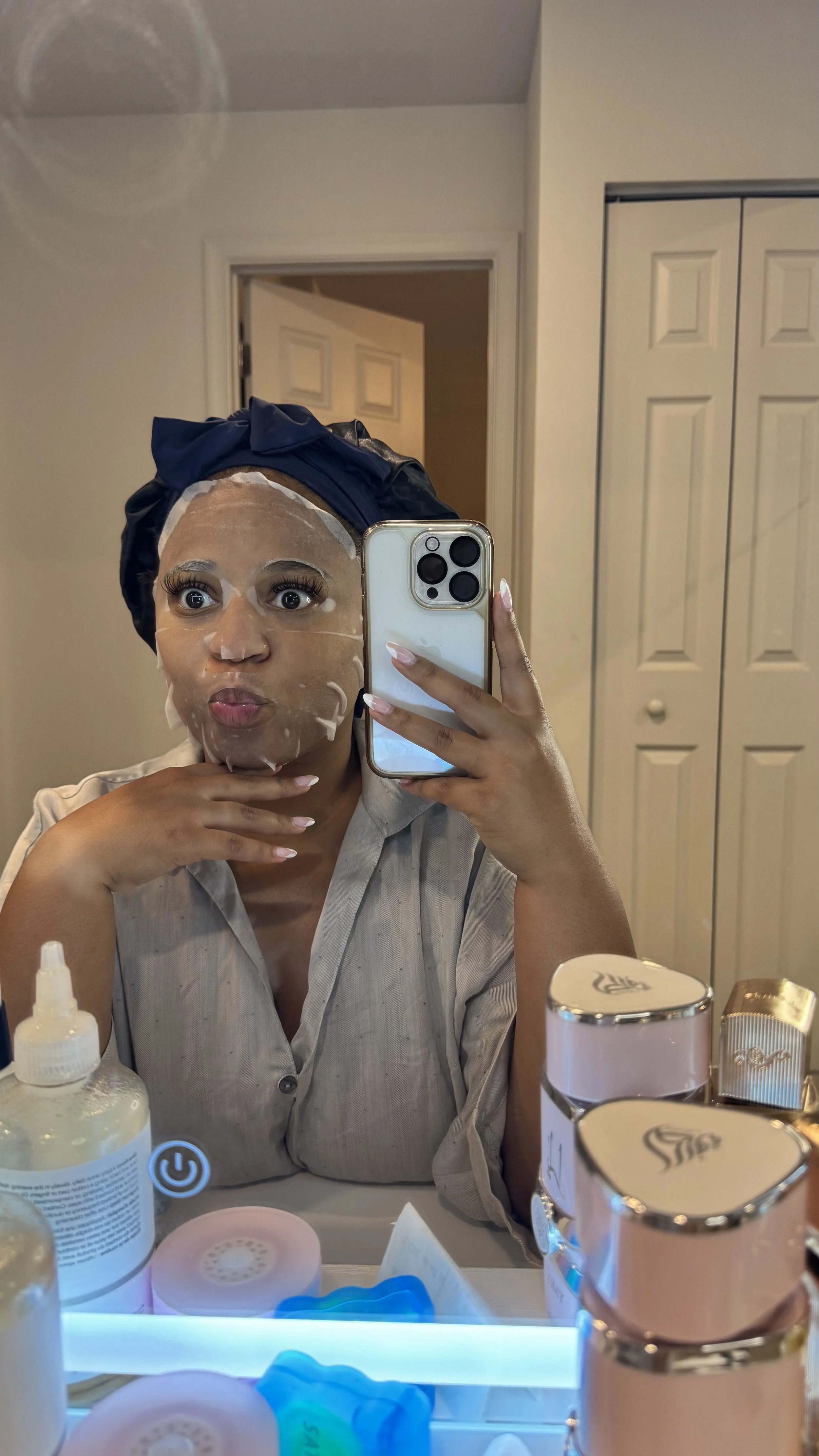 A woman in a beige top taking a mirror selfie with a phone, wearing a blue hair wrap, and a sheet mask on her face, in a bathroom with skincare products and a closet in the background.