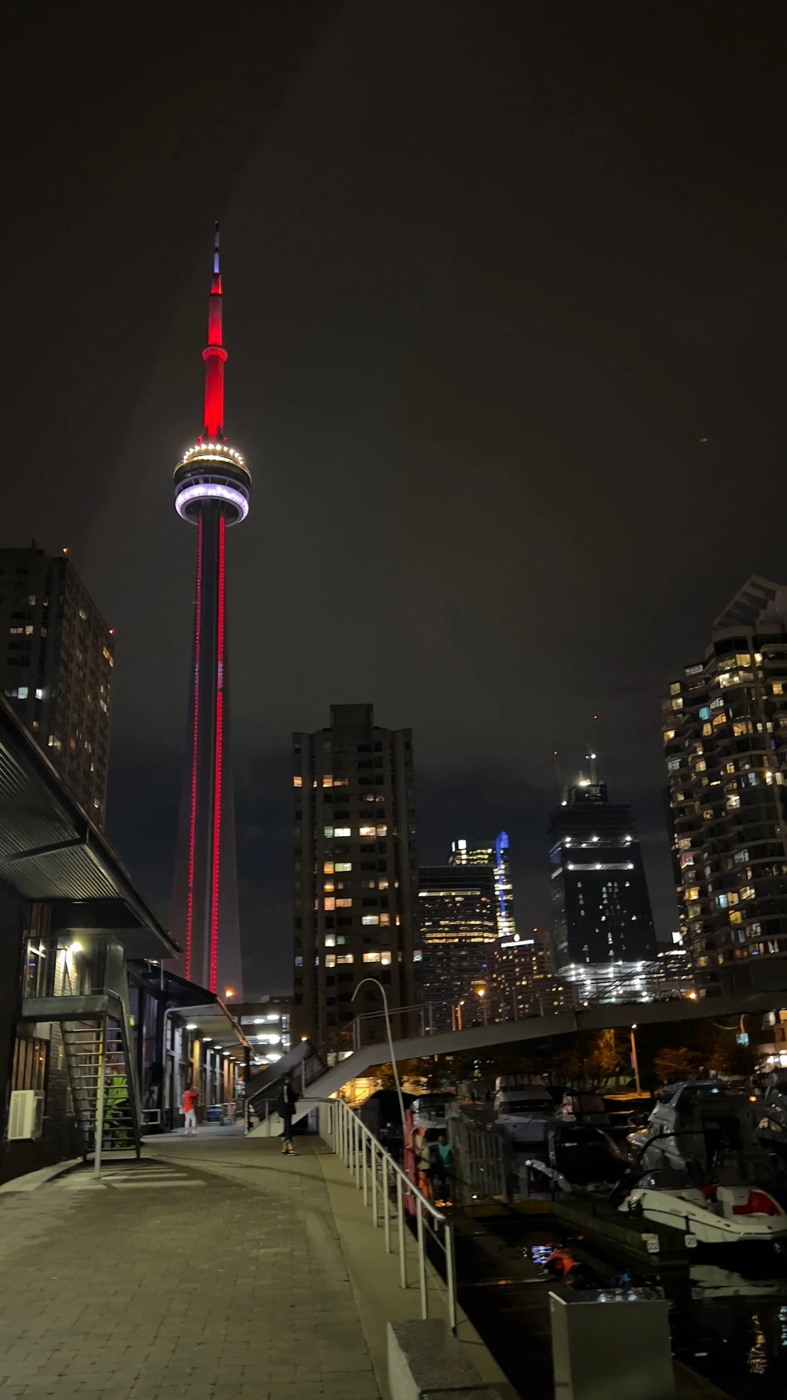 Night view of Toronto skyline with the CN Tower illuminated in red, surrounded by tall buildings and a marina with boats.