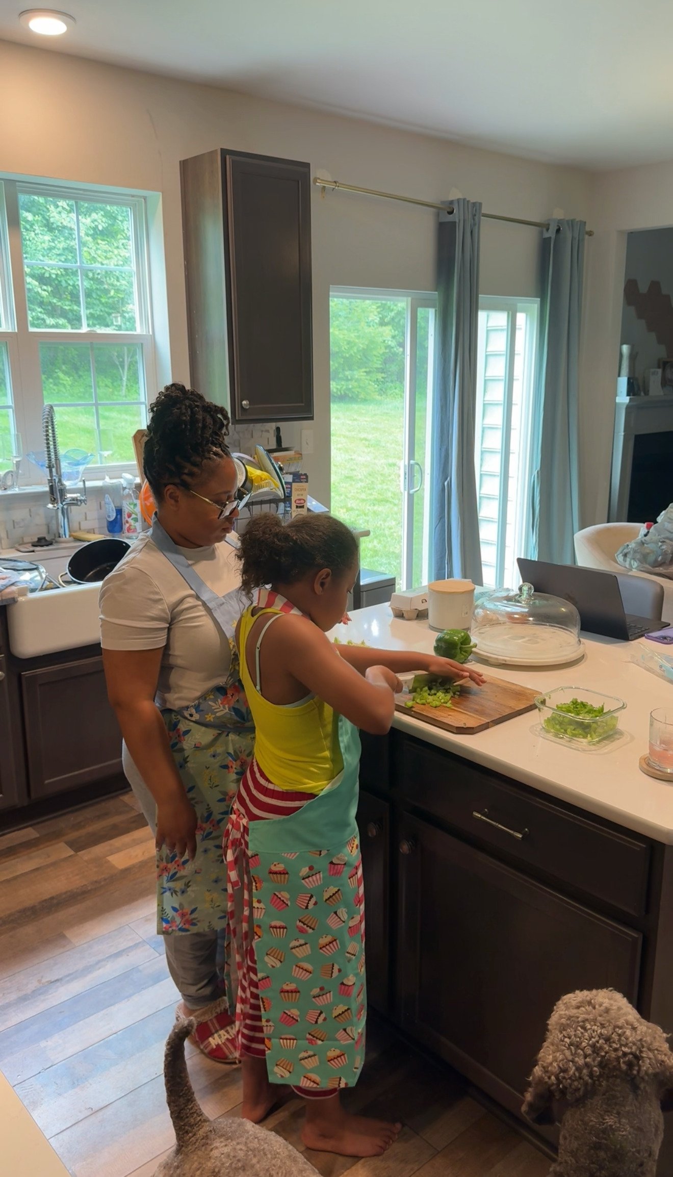 A woman and a young girl chopping celery together in a kitchen. The woman is standing behind the girl, guiding her. The kitchen has dark cabinets, a white countertop, and a window behind them showing green grass outside.