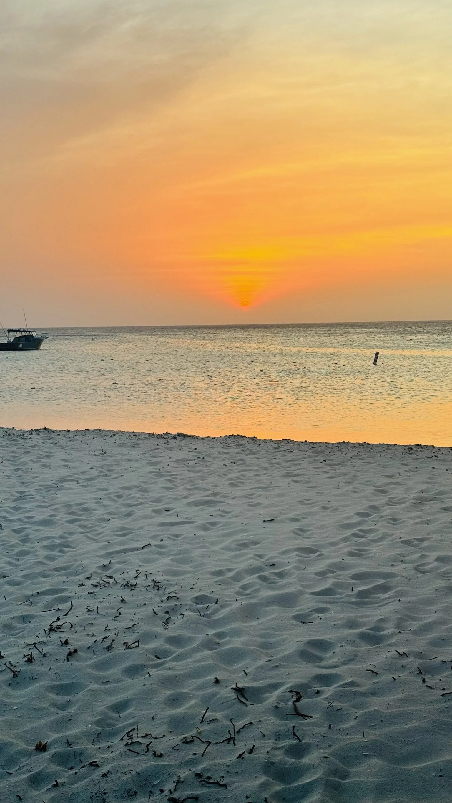 Aruba beach with sandy shore, calm waters, a boat on the left, and a vibrant sunset with the sun just above the horizon in the background.