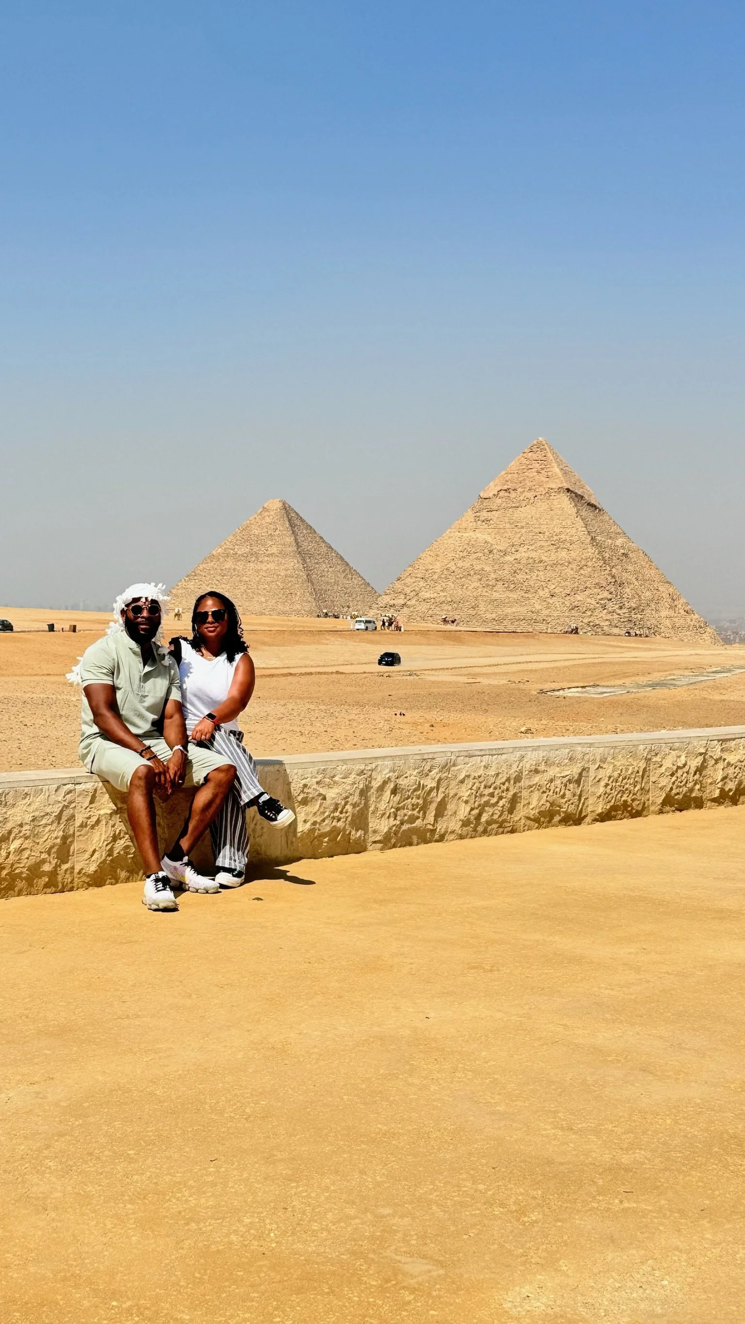 Two people sitting in front of the pyramids at Giza, Egypt, with a clear blue sky overhead.