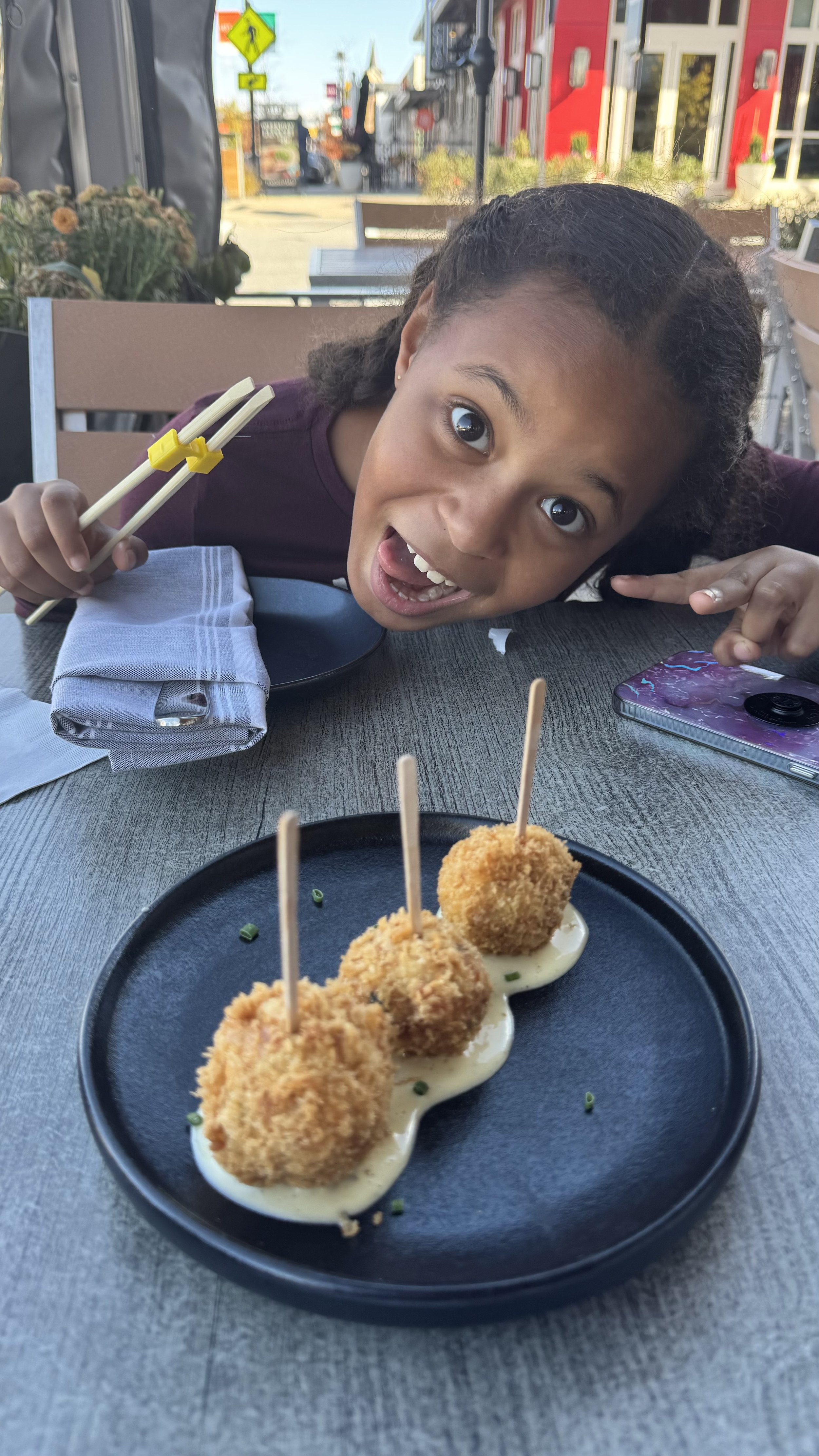 A young girl with curly hair excitedly poses at an outdoor restaurant table with a plate of three breaded and fried appetizers on a bed of white sauce.