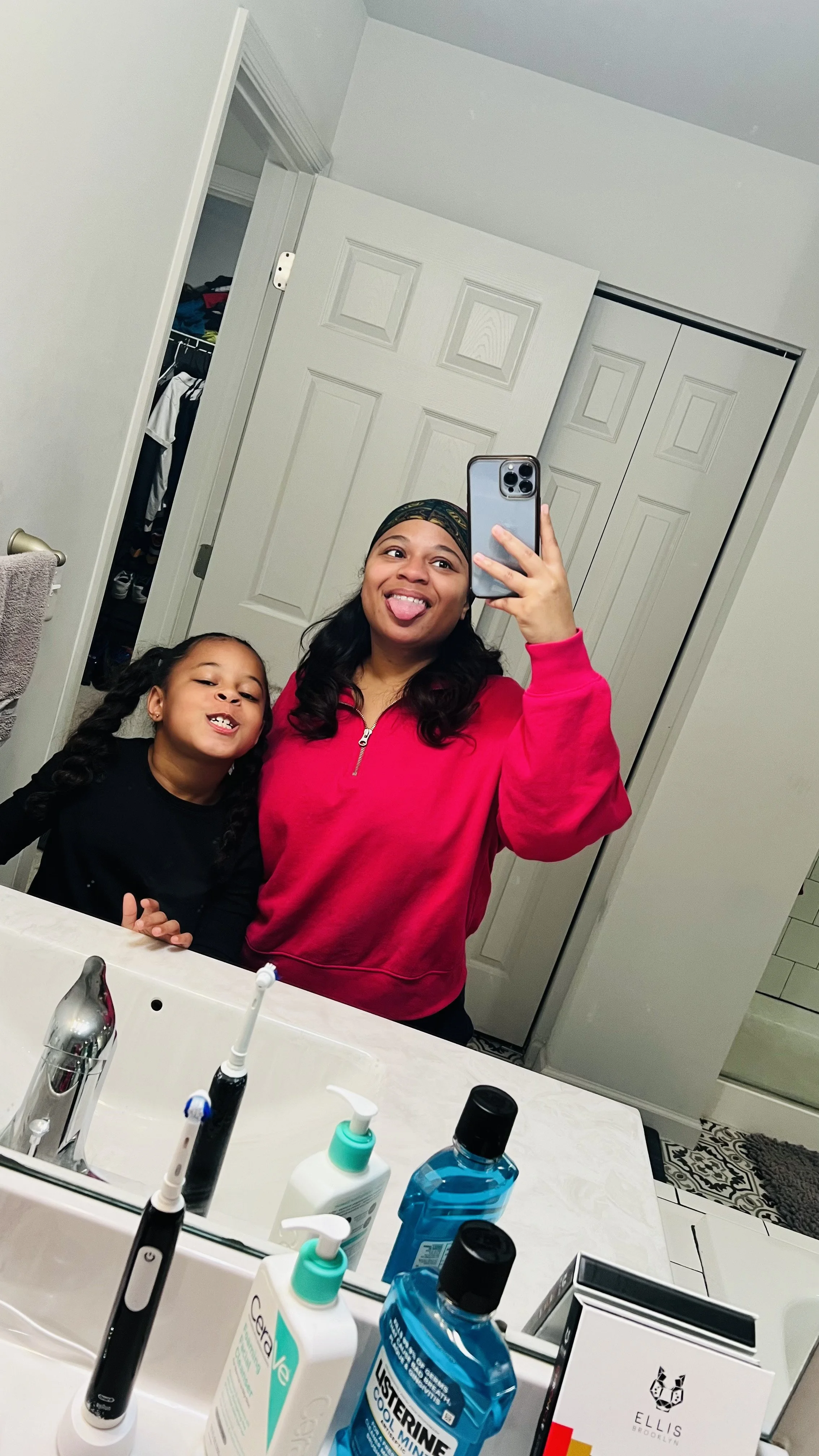 A woman and a young girl are taking a mirror selfie in a bathroom. The woman is wearing a pink sweatshirt and a headscarf, sticking out her tongue. The girl is smiling with her mouth slightly open. The bathroom countertop has various toiletries, incl